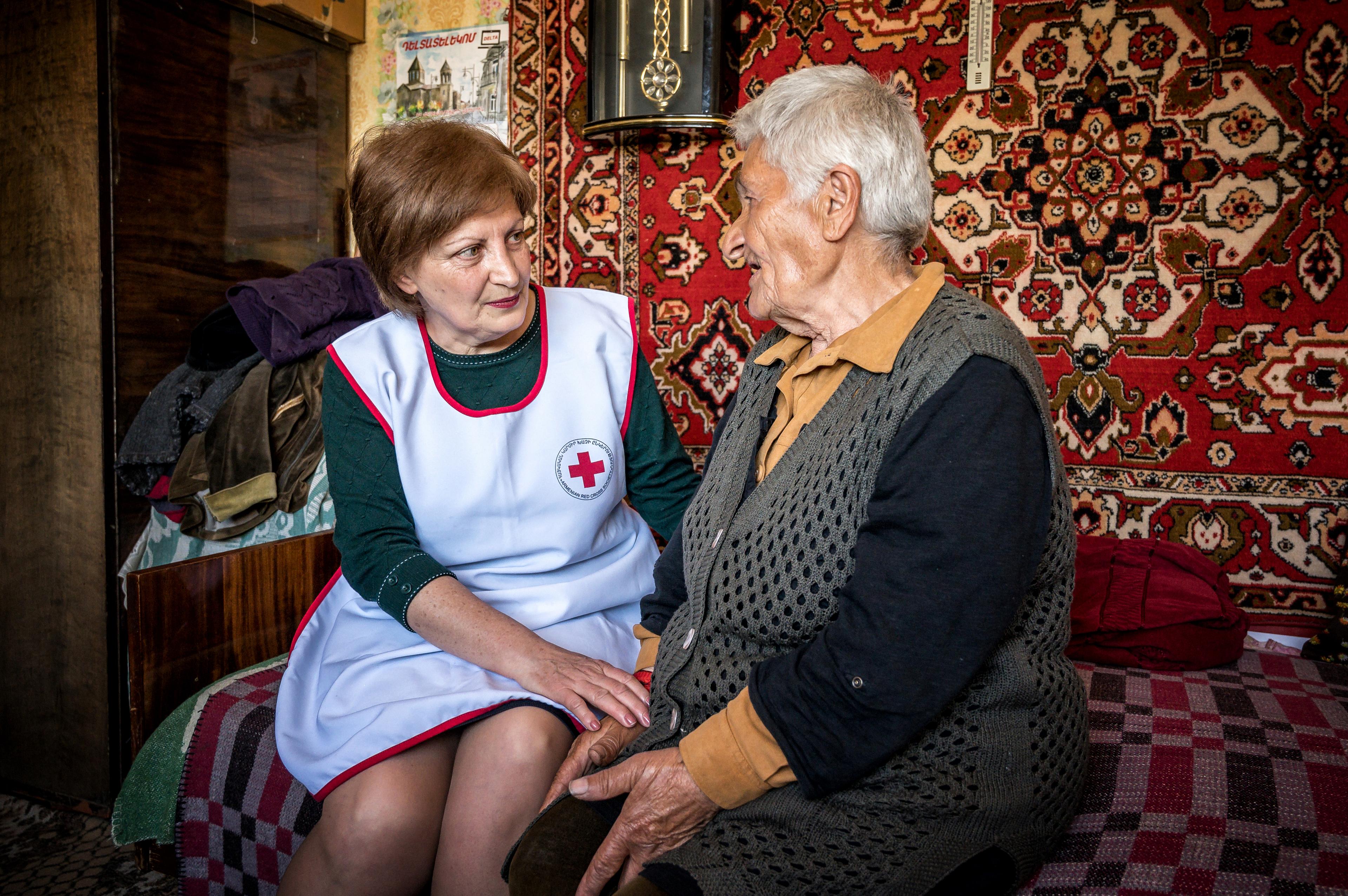 Une femme portant un gilet de la Croix-Rouge est assise en face d'une dame âgée. Elles sont assises sur un lit et des tapis sont accrochés au mur derrière elles.