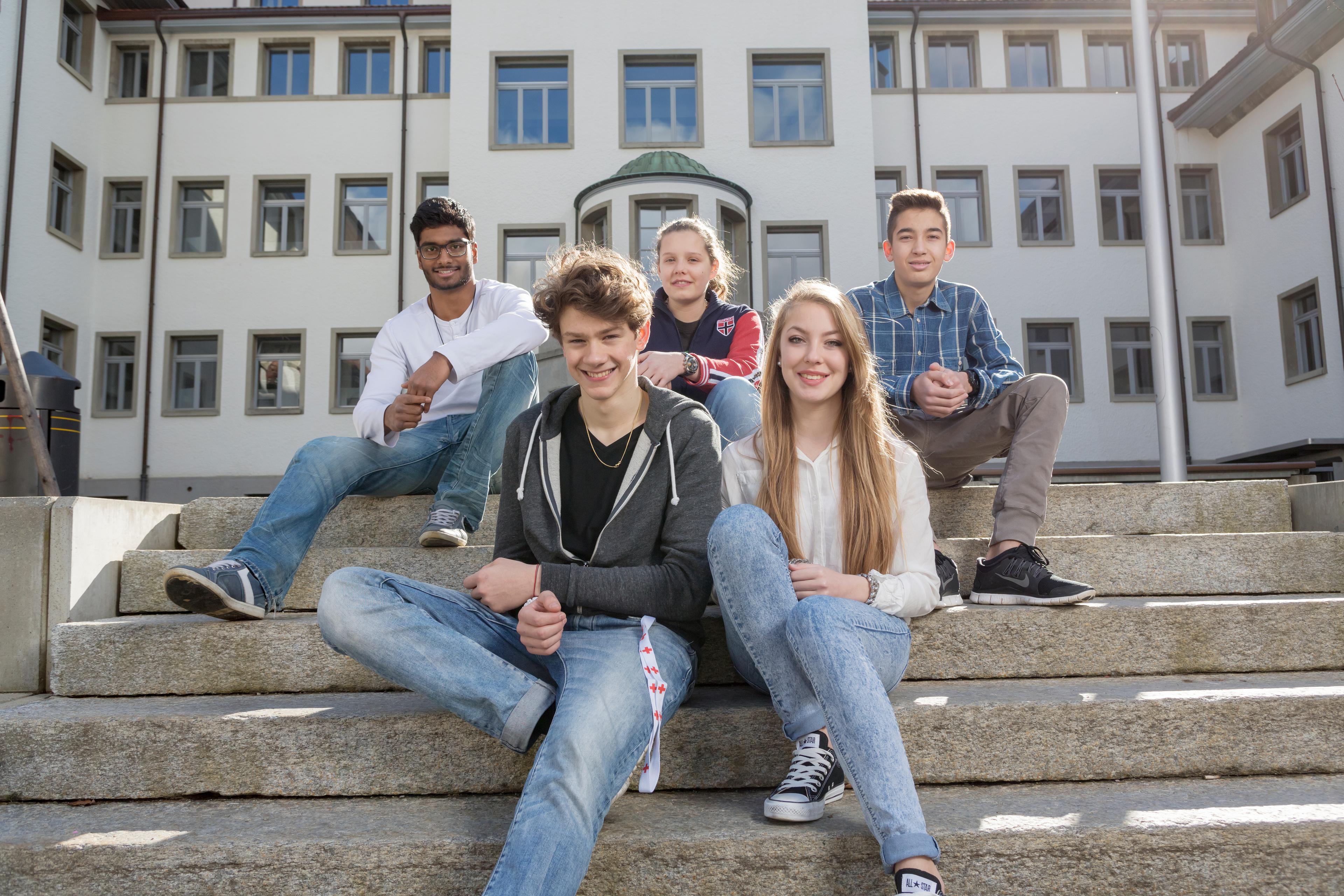 Cinq jeunes gens sur un escalier, un bâtiment scolaire en arrière-plan. Ils sourient gentiment.