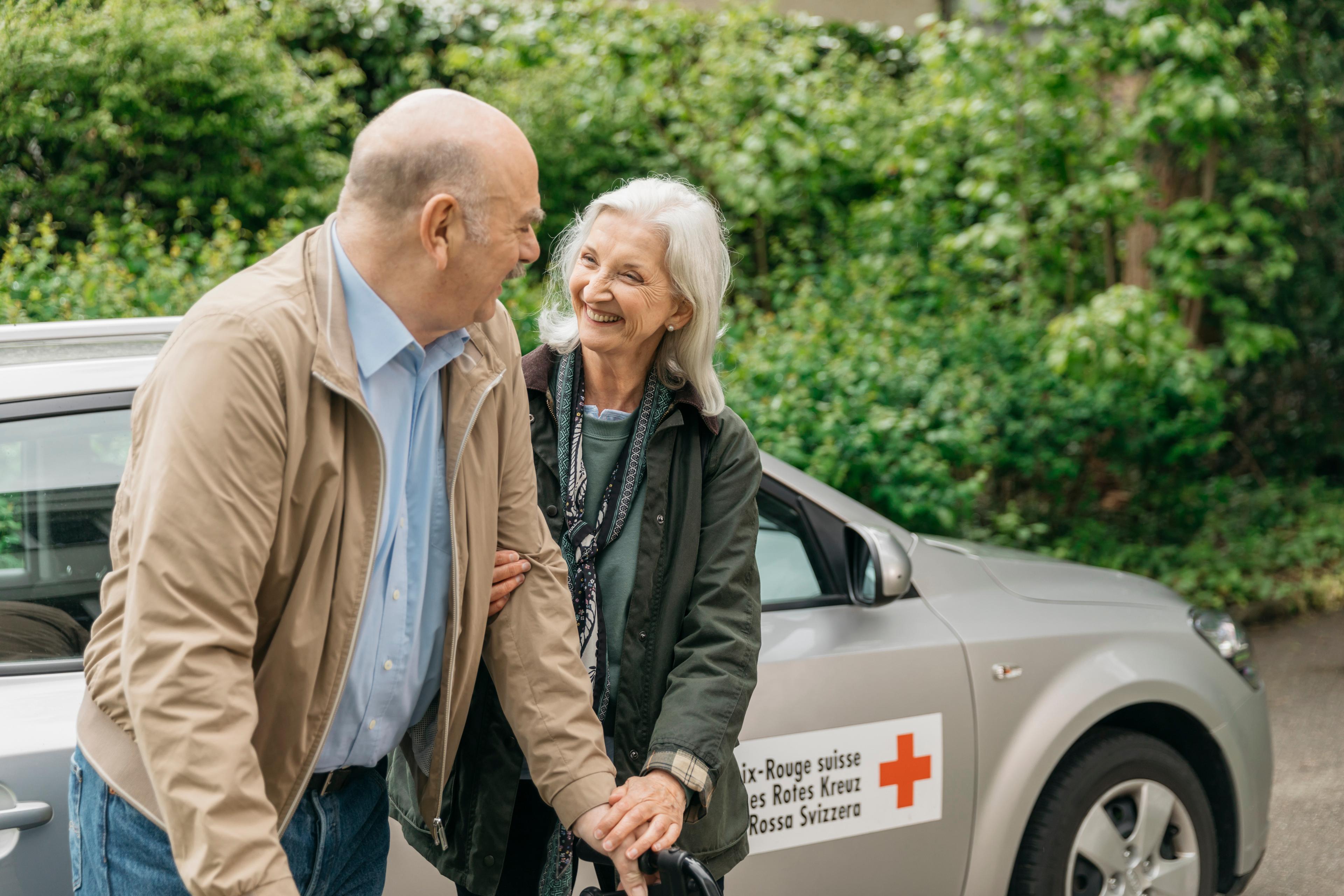 Eine freiwillige Fahrerin stützt den Arm eines Mannes mit eingeschränkter Mobilität. Sie stehen vor einem Fahrzeug des Schweizerischen Roten Kreuzes und lächeln sich an.