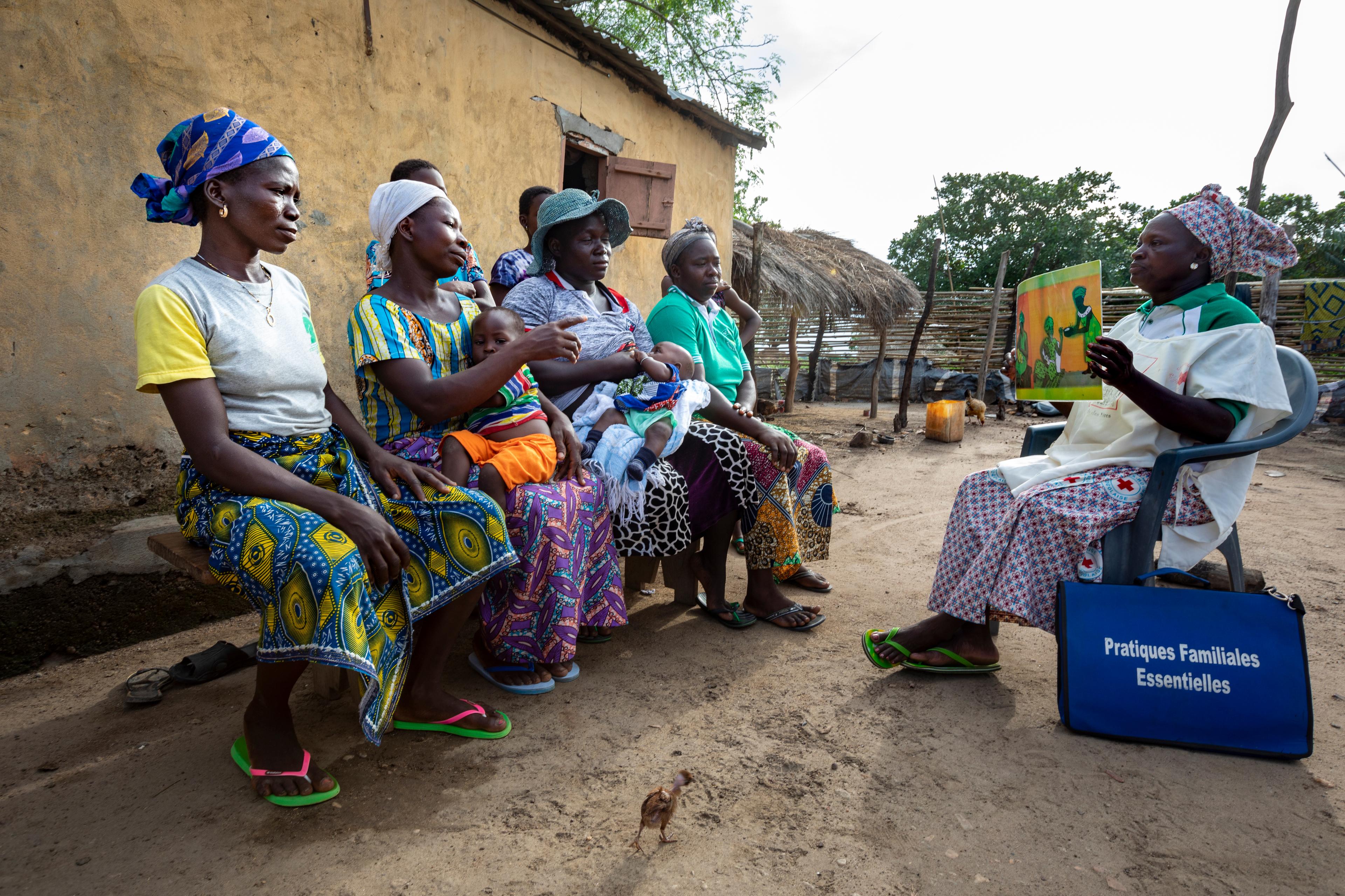 Four African women sit on a wooden bench in front of a mud hut. Two are carrying babies. Opposite them sits a woman wearing a Red Cross vest. She shows the women an illustration of a village scene with mothers and other women.