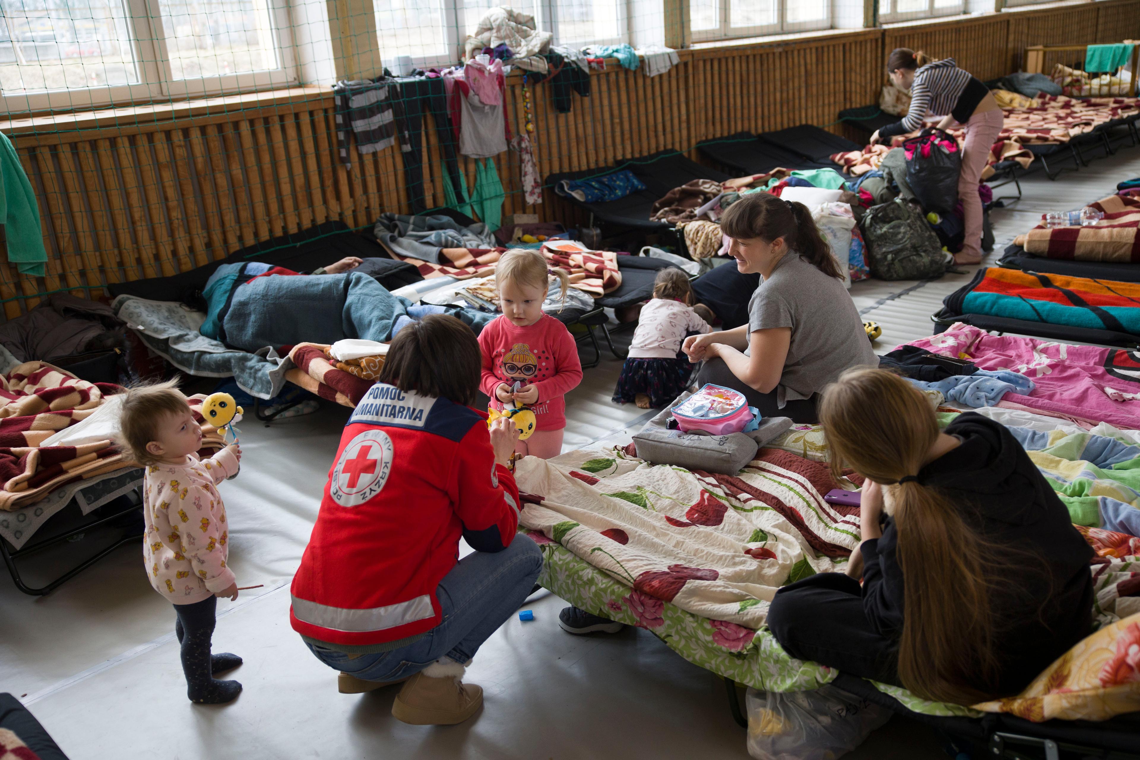 Eine Frau mit Rotkreuz-Jacke kauert neben zwei jungen Frauen und ihren kleinen Kindern. Sie befinden sich in einer Notunterkunft mit Dutzenden von aneinandergereihten Betten und Matratzen.