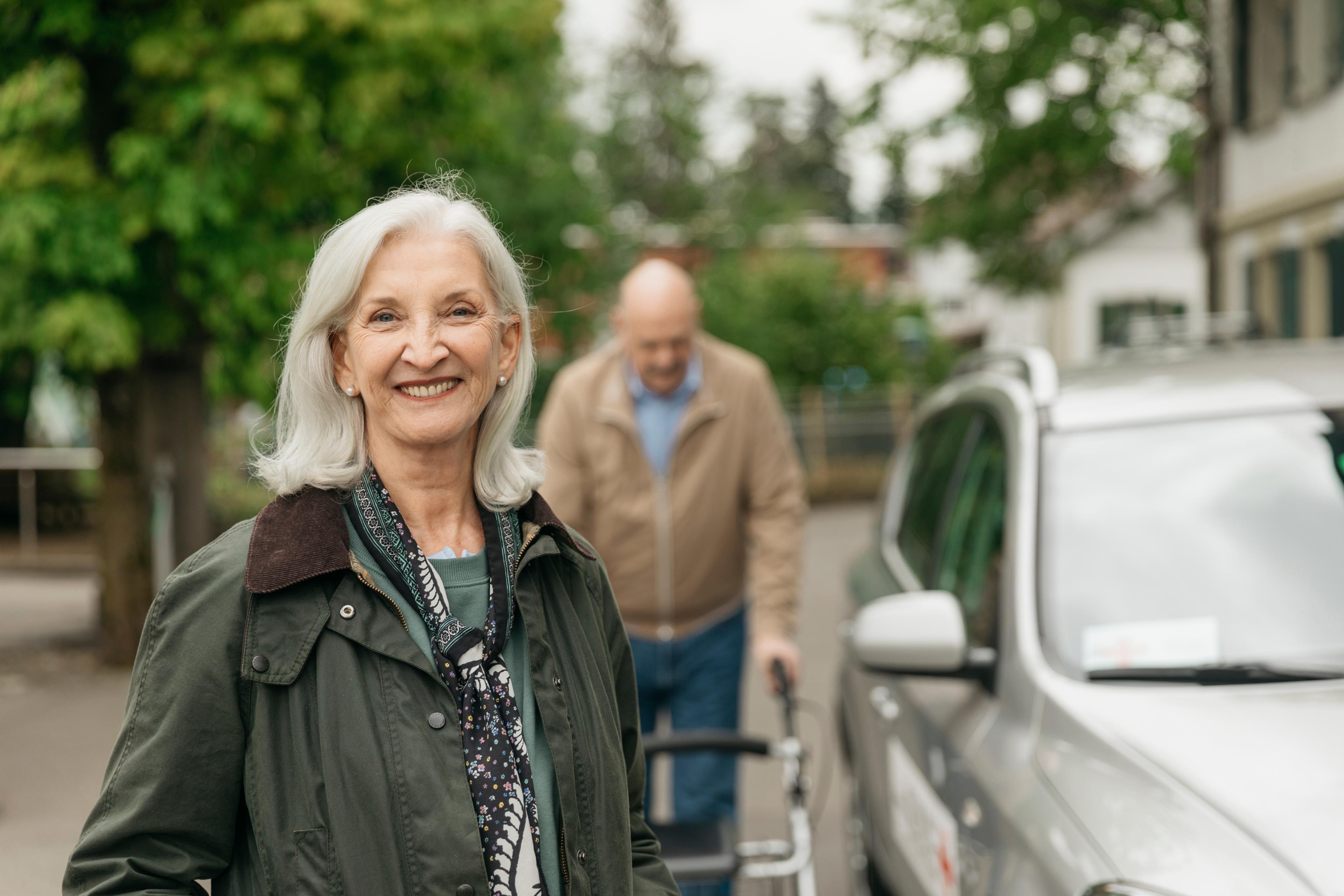 Transport service: A woman is smiling into the camera. Behind her is an elderly man with a rolling walker.