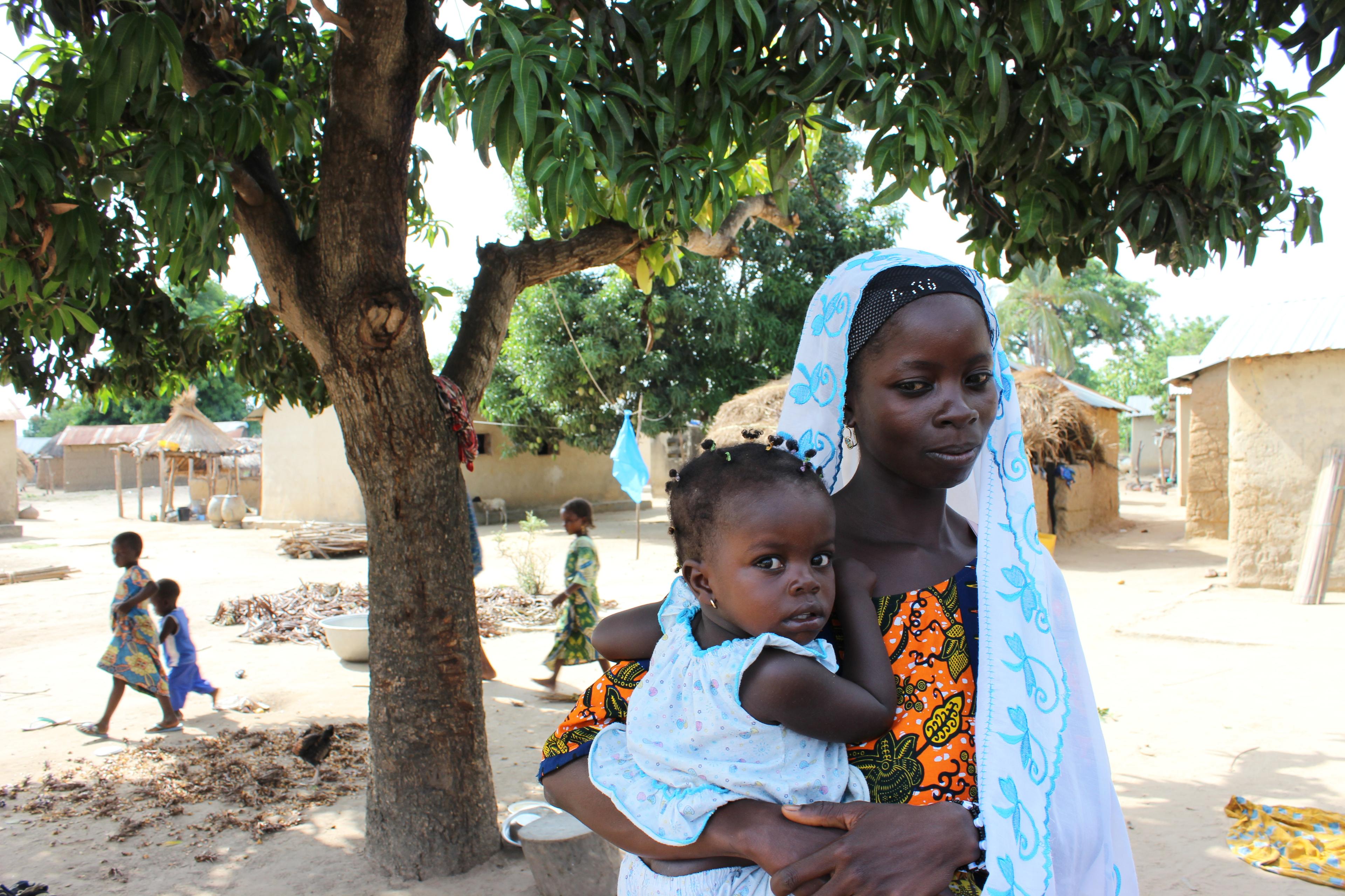 A mother holds an infant in her arms. They are standing in a village under a tree in the shade. In the background there are other children.