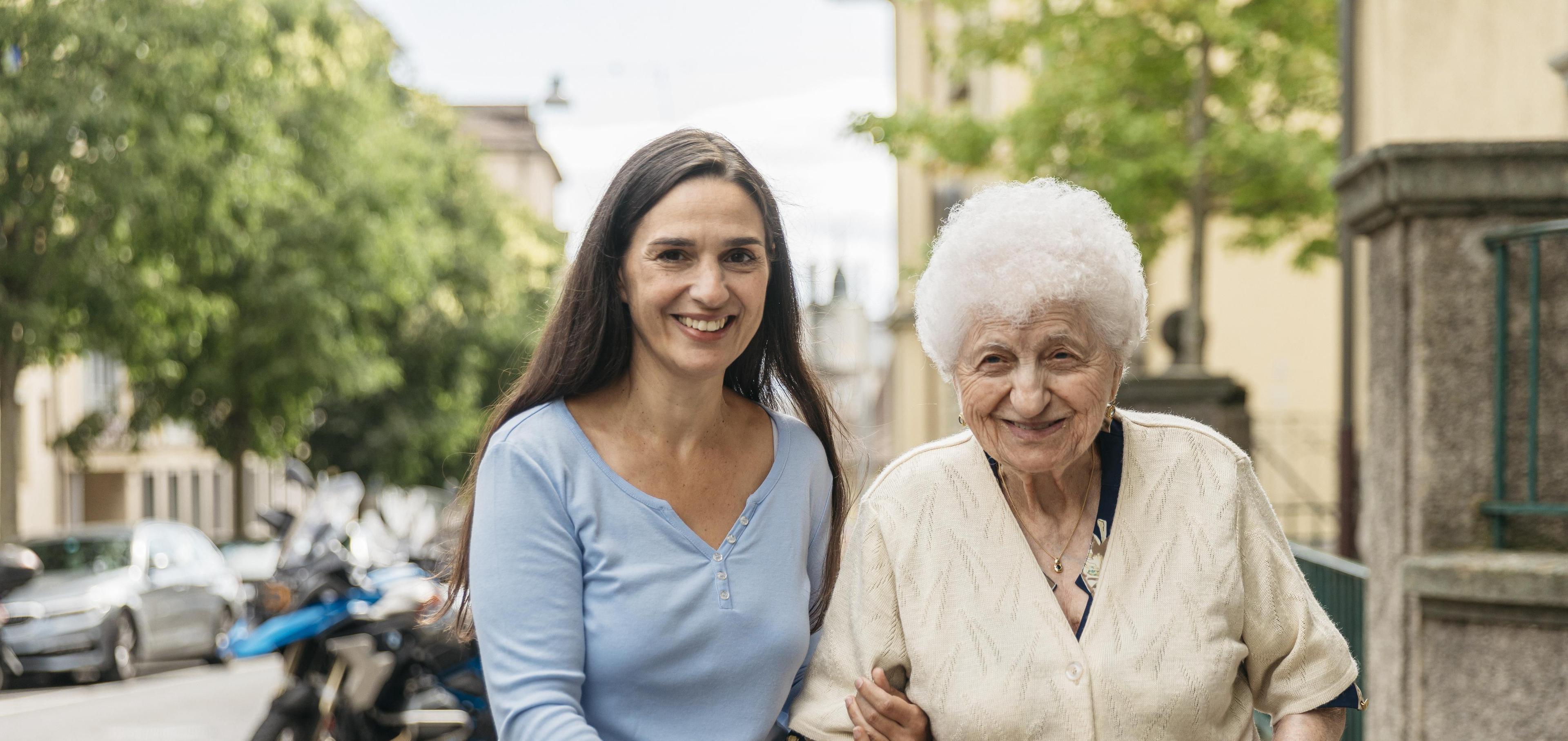 Eine jüngere Frau Arm in Arm mit einer älteren Dame beim Spaziergang.