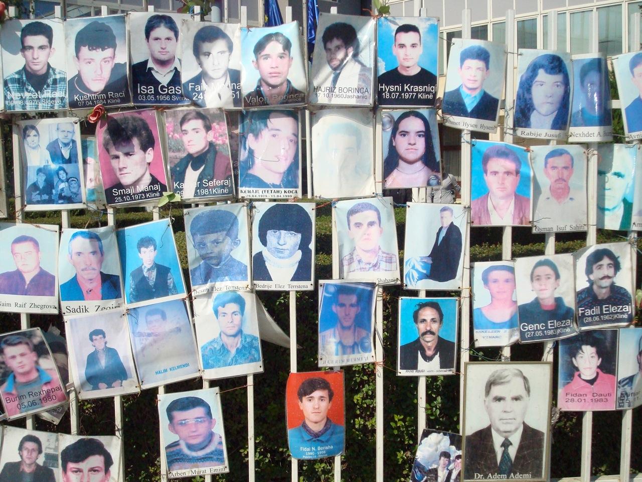 A large number of portrait photos are hung on a fence. They are missing persons. The names and sometimes the date of birth are written on the photos.
