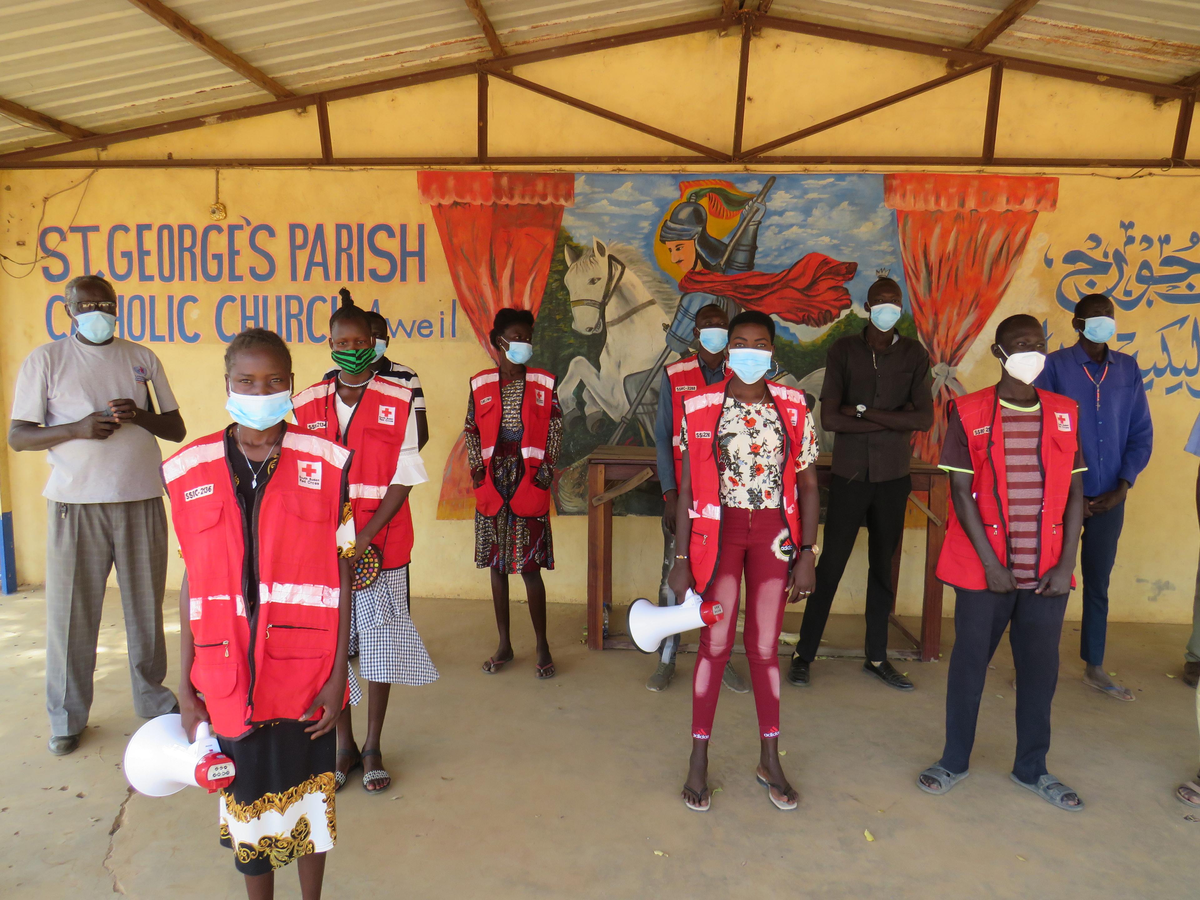 Auf dem Bild sind Rotkreuz-Freiwillige, die voneinander Abstand haben und in die Kamera schauen in einem Gebäude zu sehen. Sechs von ihnen tragen eine rote Weste mit dem Emblem des Roten Kreuzes.