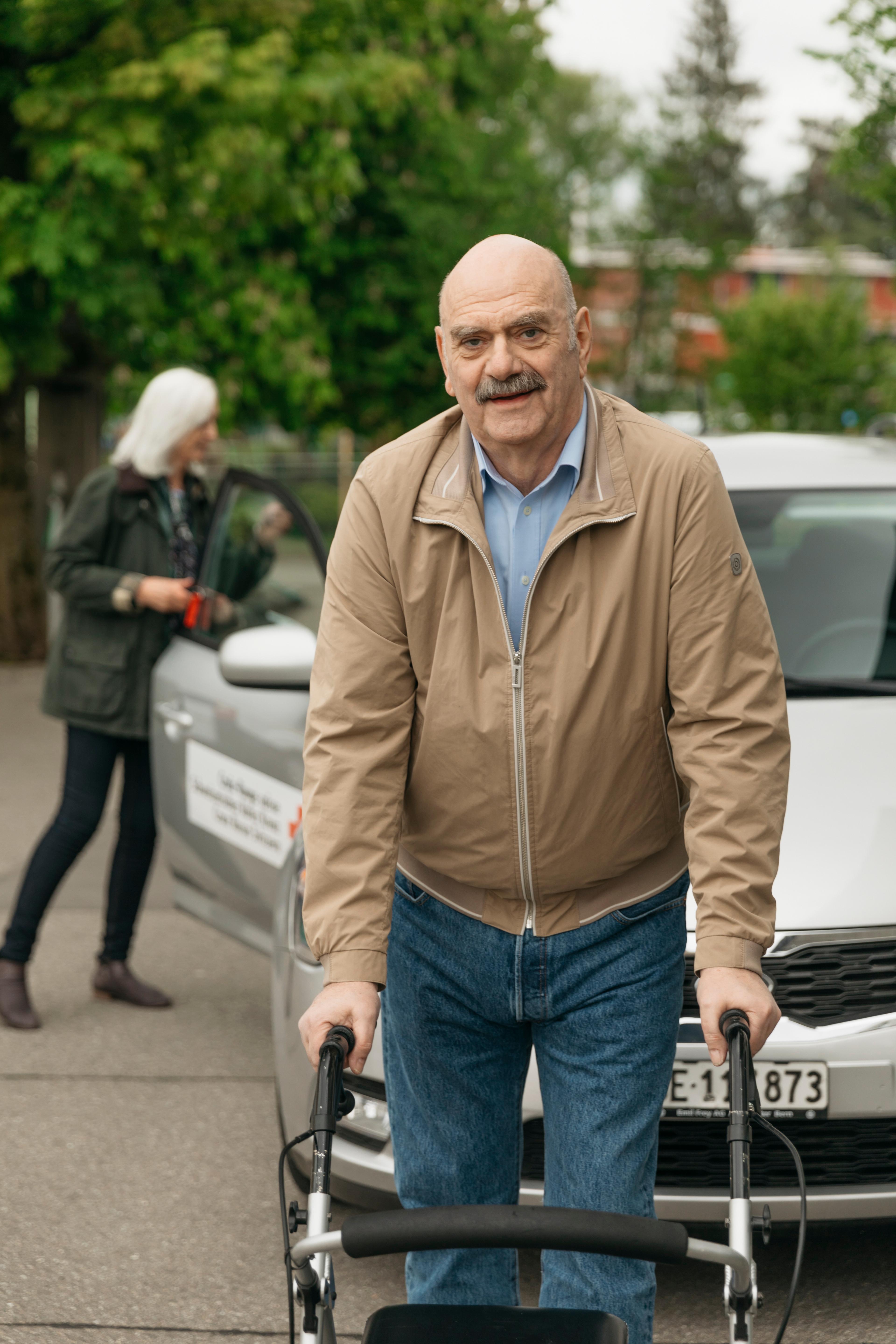 Service des transports: un homme âgé appuyé sur son déambulateur se tient devant une voiture et fixe l’objectif en souriant.