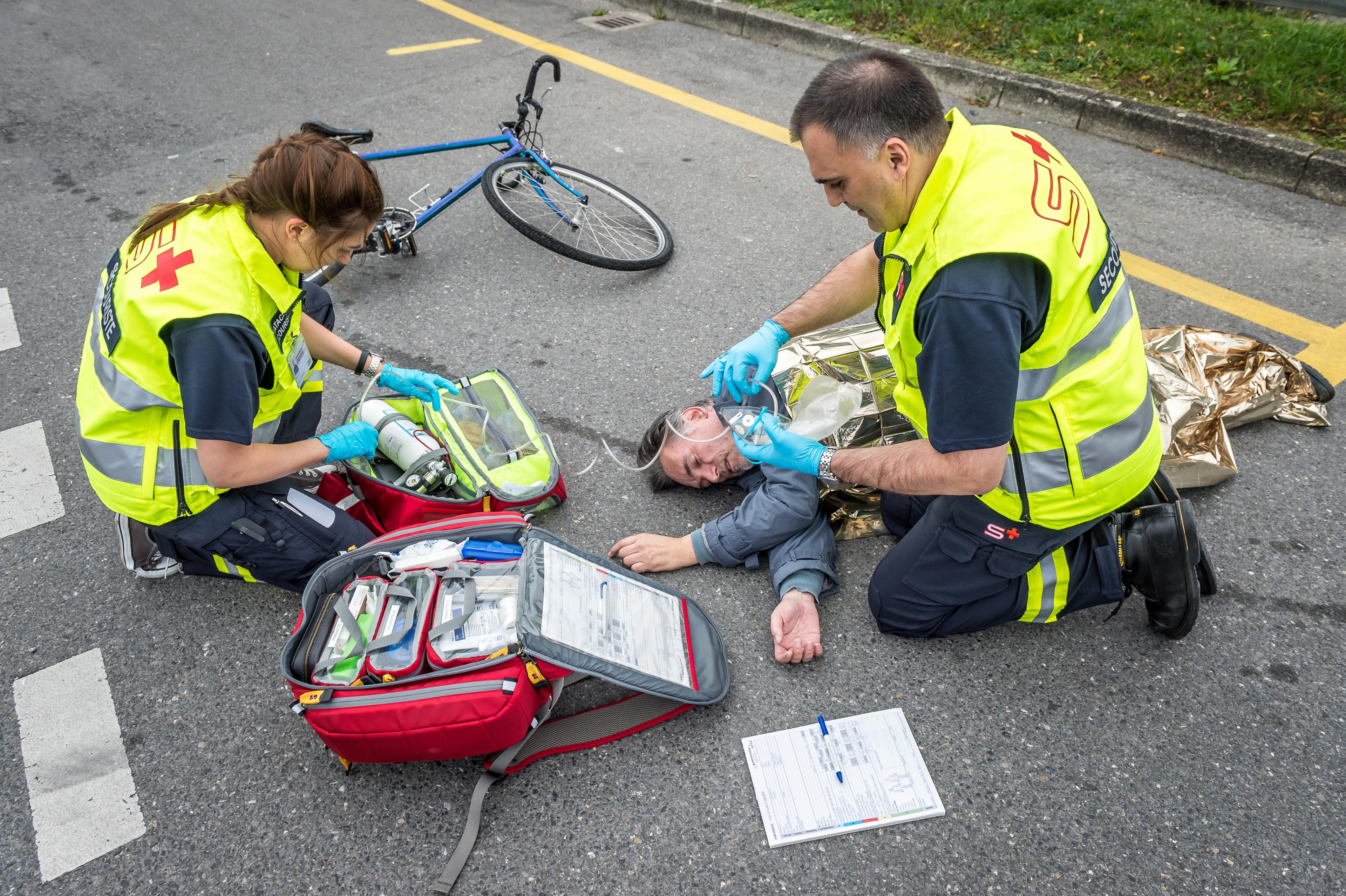 Esercizio del samaritano: un ciclista ferito giace sulla strada accanto alla sua bicicletta, due samaritani si prendono cura di lui.