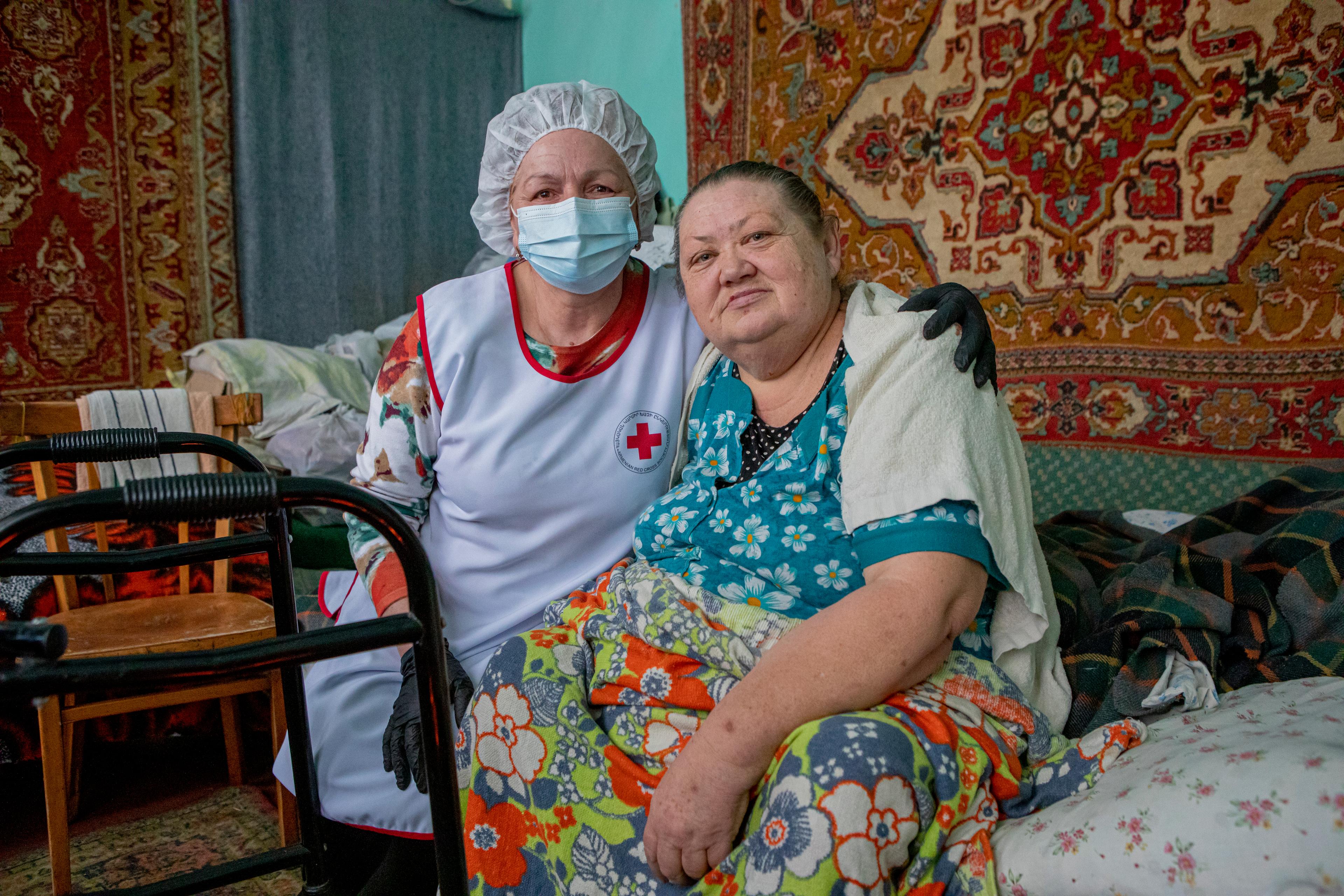 Eine Rotkreuz-Pflegerin sitzt neben einer älteren Frau in ihrem Bett. Im Hintergrund sieht man Teppiche an der Wand.