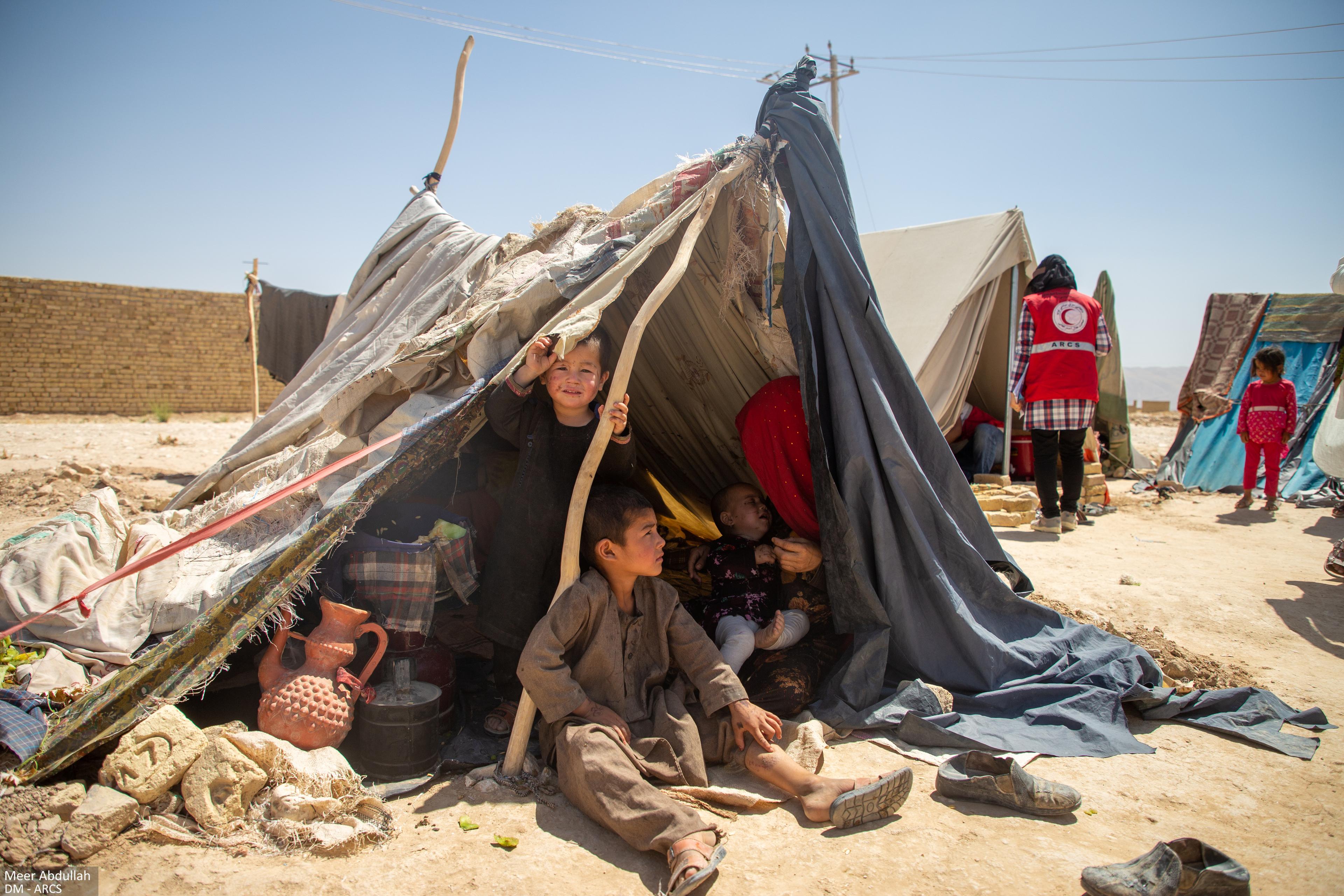 Drei Kinder und eine Frau in einem selbstgebastelten Zelt in Afghanistan. Im Hintergrund sind ähnliche Zelte zu sehen.