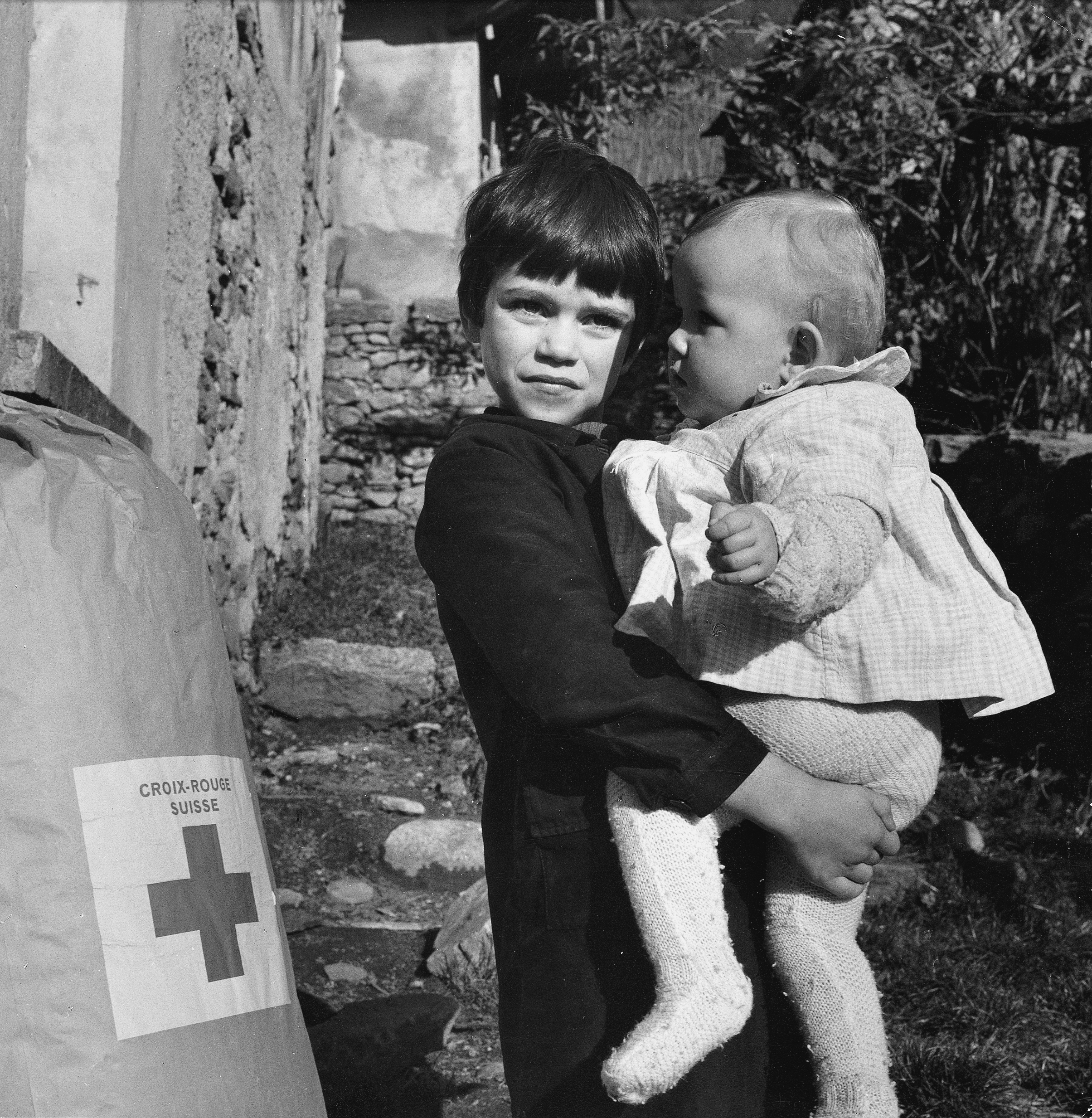 Une photo en noir et blanc d'un enfant avec un petit enfant dans les bras, à côté d'un sac de biens de secours avec l'emblème de la. CRS