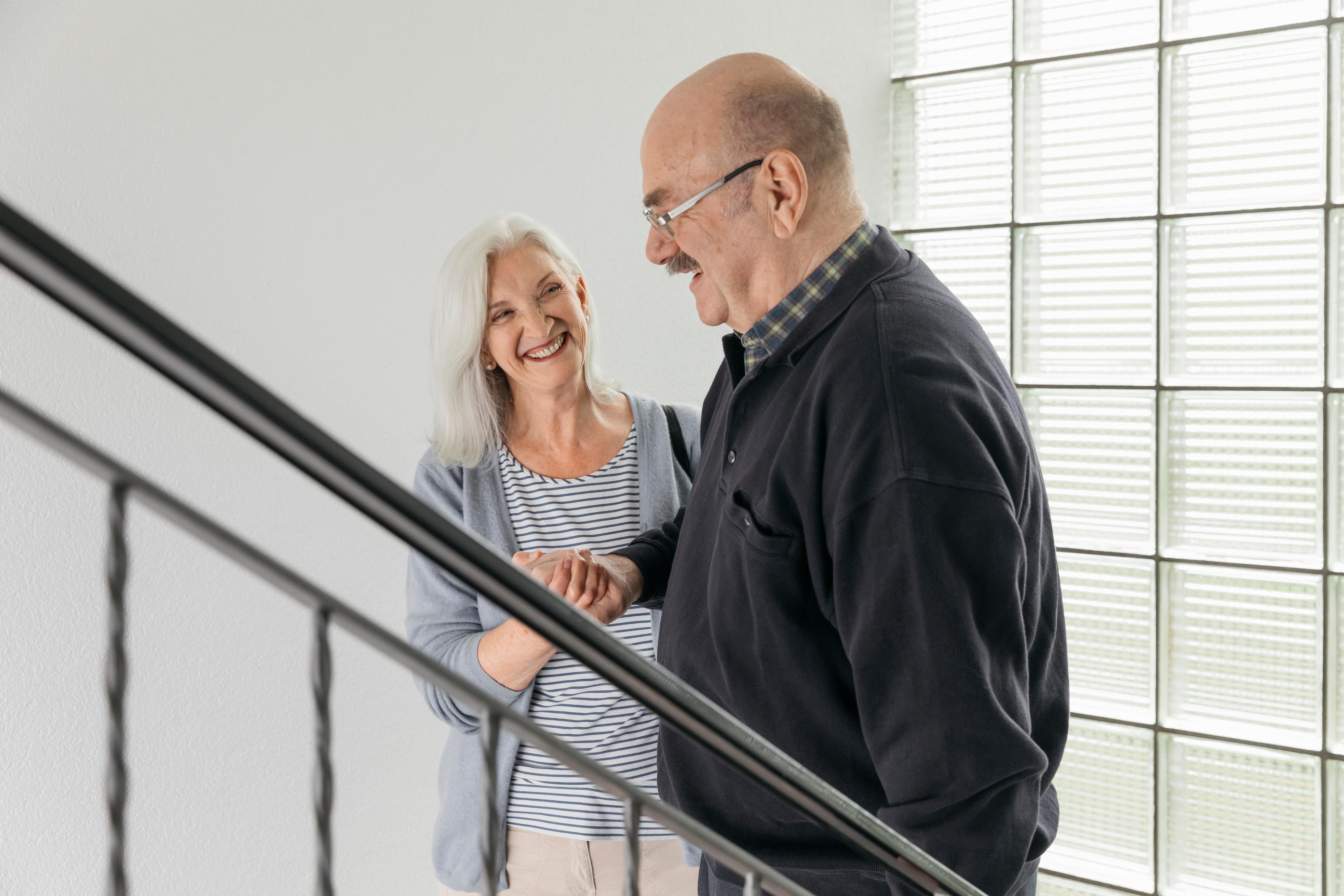 Fahrdienst: Eine ältere Frau und ein älterer Herr stehen im Treppenhaus und gehen die Treppe herunter. Die Frau stütz den Mann am Arm. Sie lachen sich beide an.