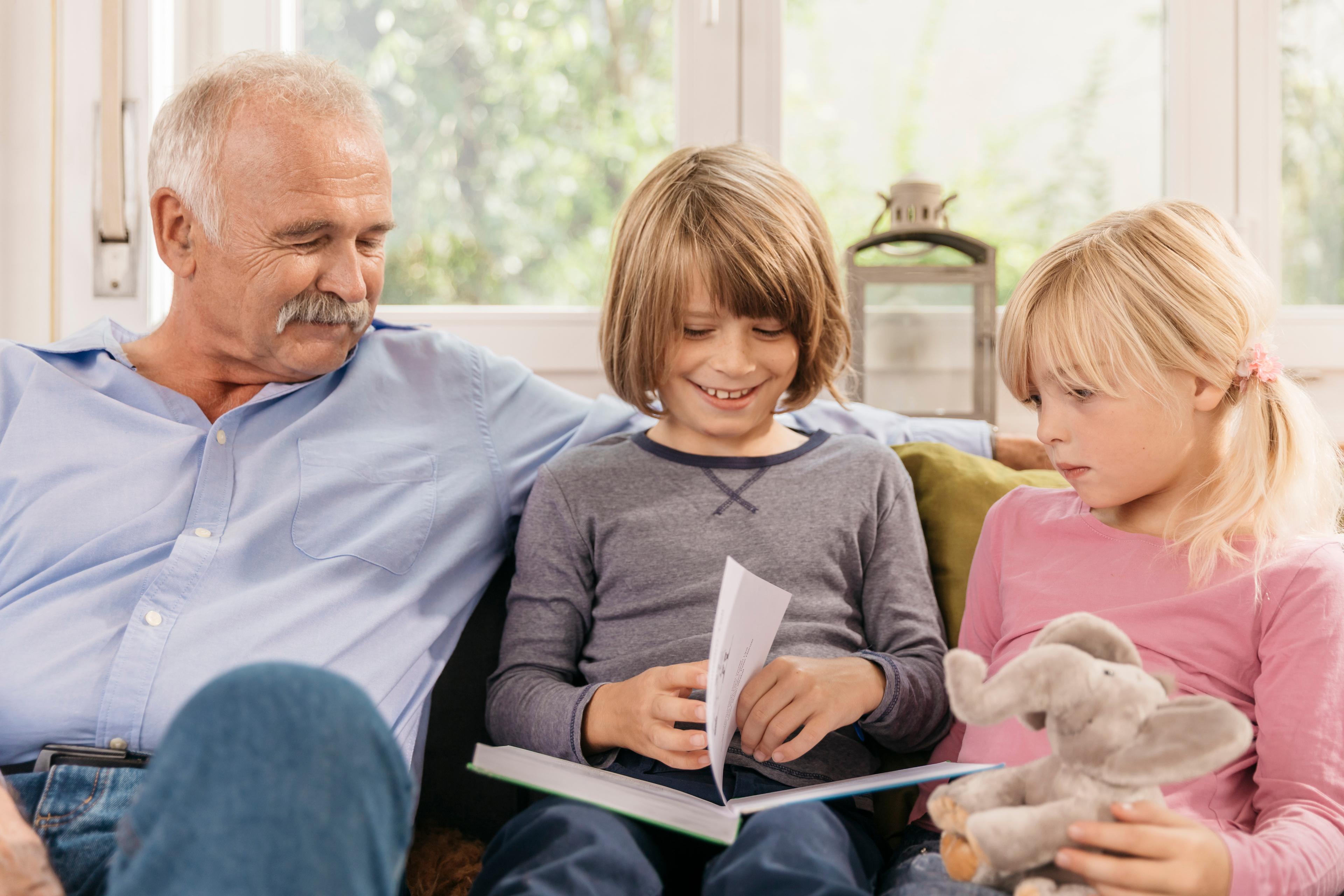 Zwei Kinder schauen sich mit ihrem Großvater auf einem Sofa ein Buch an.