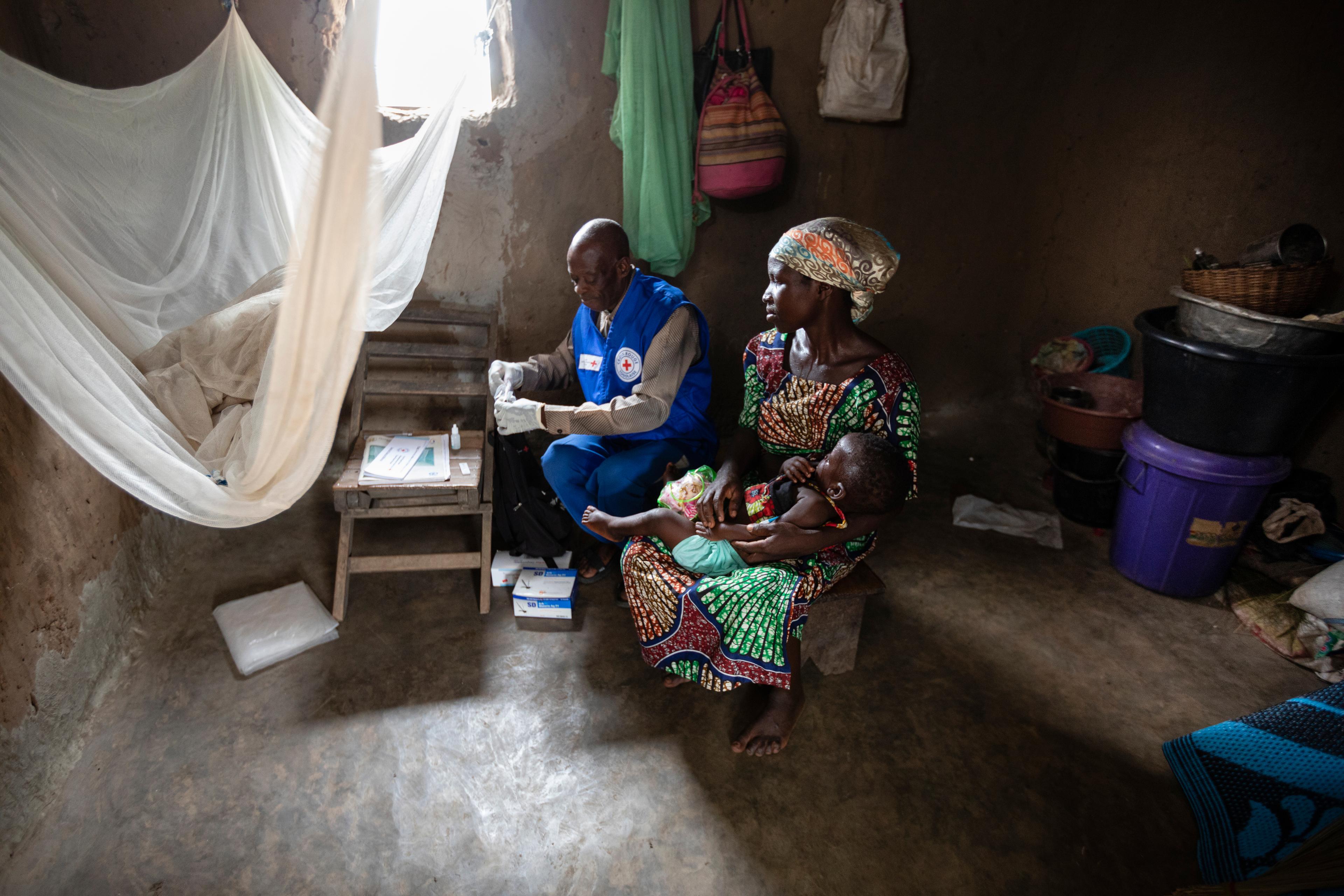 Ein Gesundheitshelfer führt bei einem Säugling, der auf dem Schoss der Mutter liegt, einen Malaria-Test durch.  