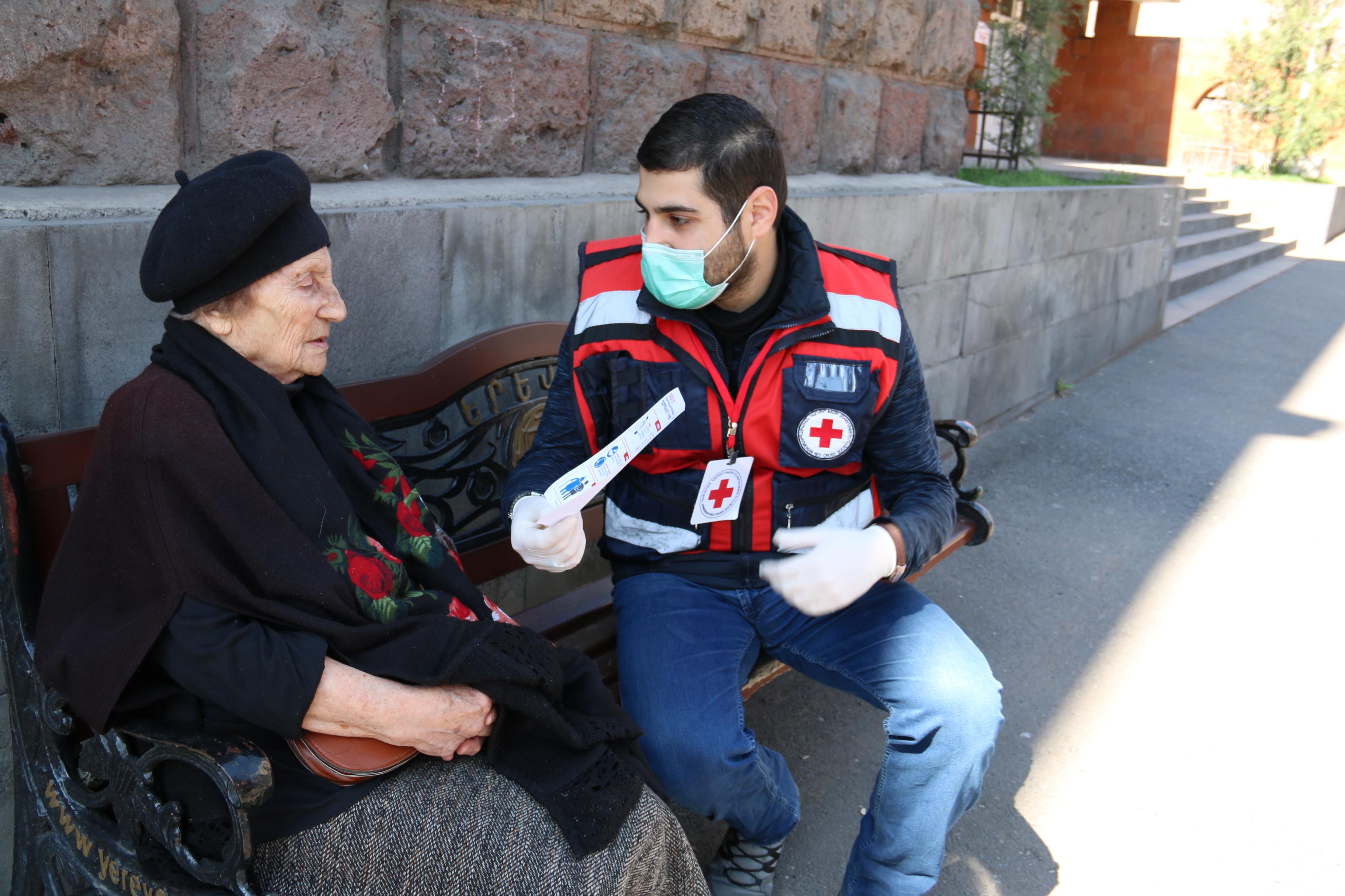 Un bénévole de la Croix-Rouge arménienne est assis sur un banc à l'extérieur avec une dame âgée et l'informe des risques de transmission du coronavirus.