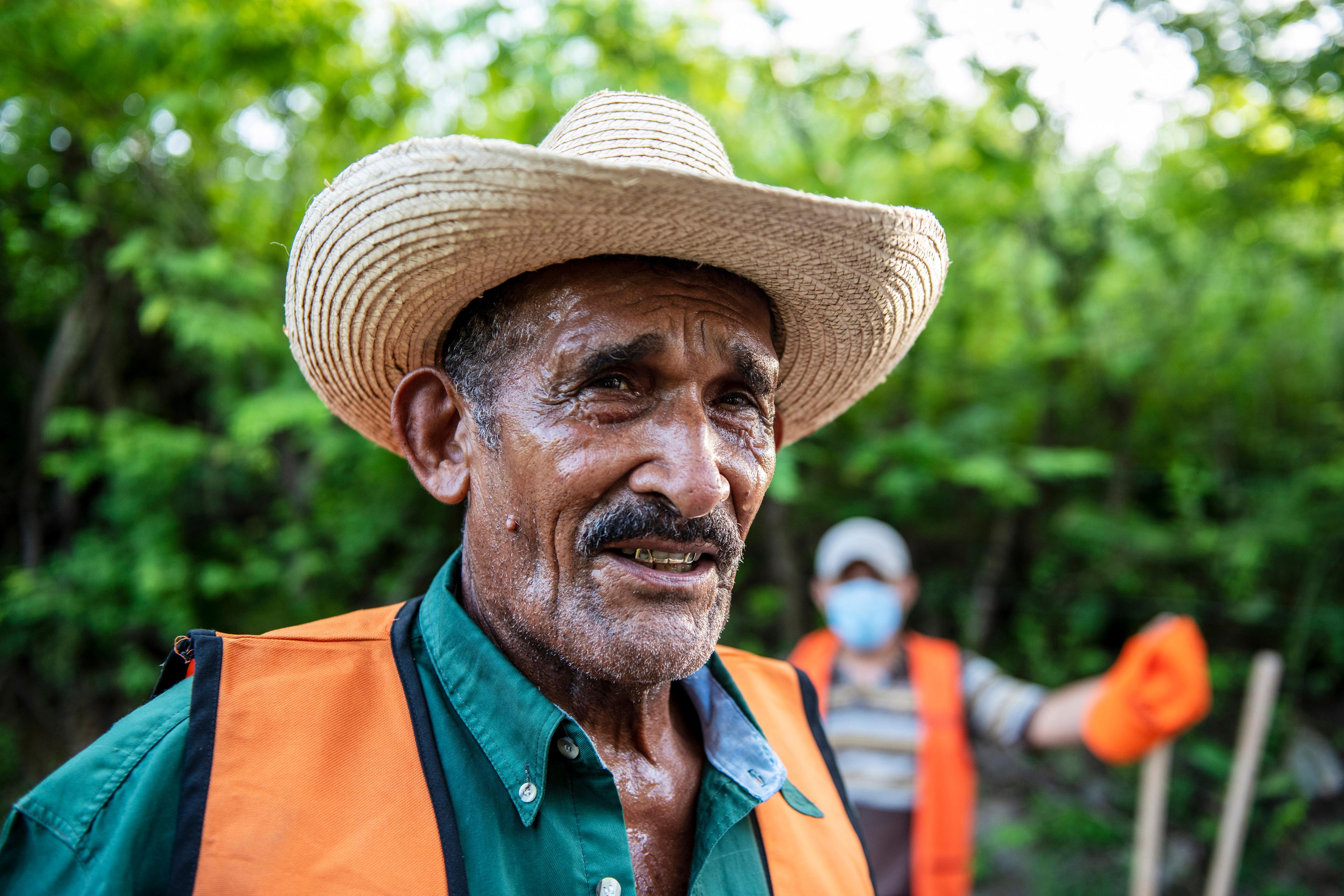 Portraitaufnahme eines älteren Mannes mit Schnauz, einem Hut und oranger Weste. Im Hintergrund befinden sich Bäume und ein weiterer Mann.