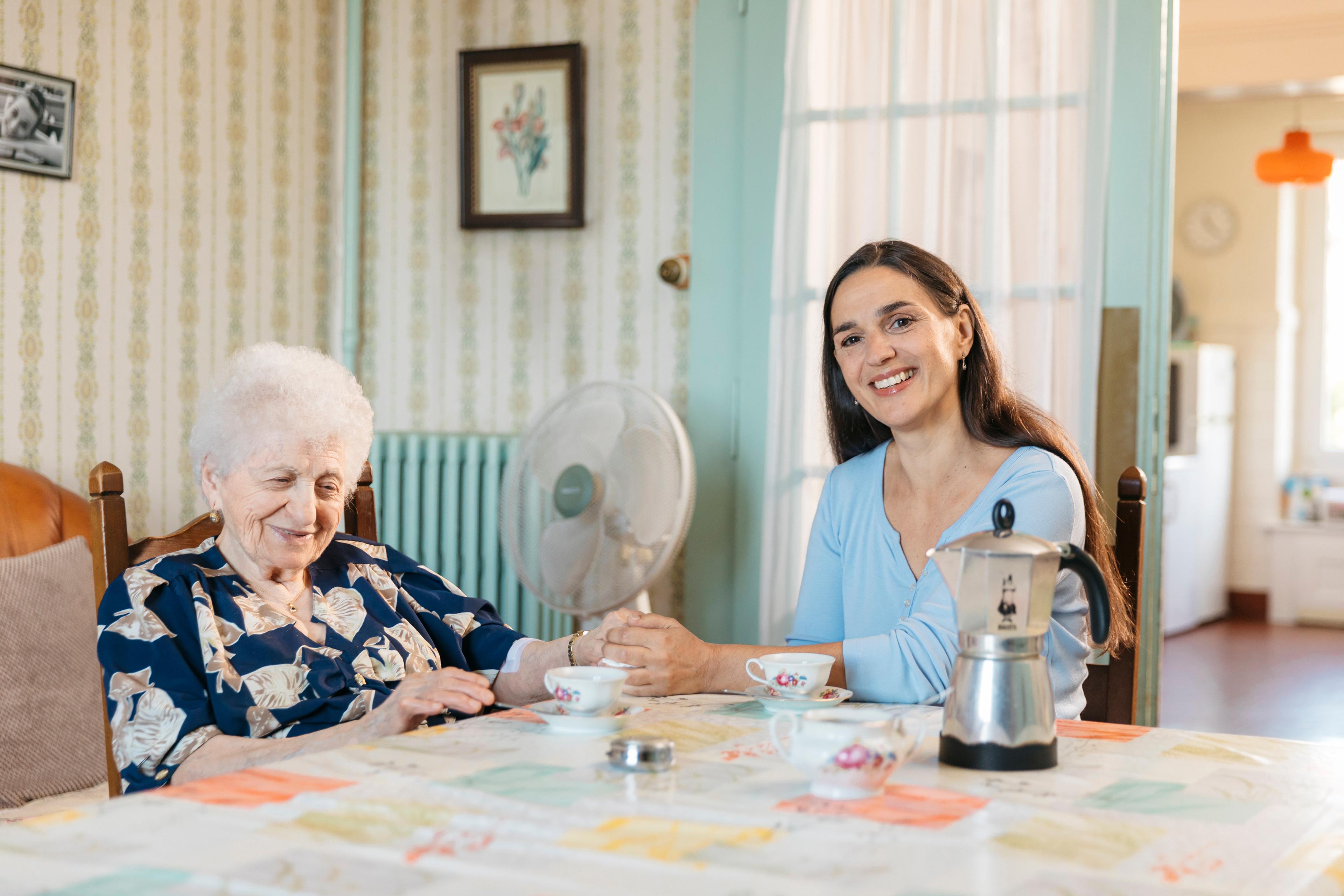 Eine Freiwillige des Roten Kreuzes sitzt mit einer älteren Dame an einem Tisch. Sie trinken zusammen Kaffee und halten sich an der Hand.