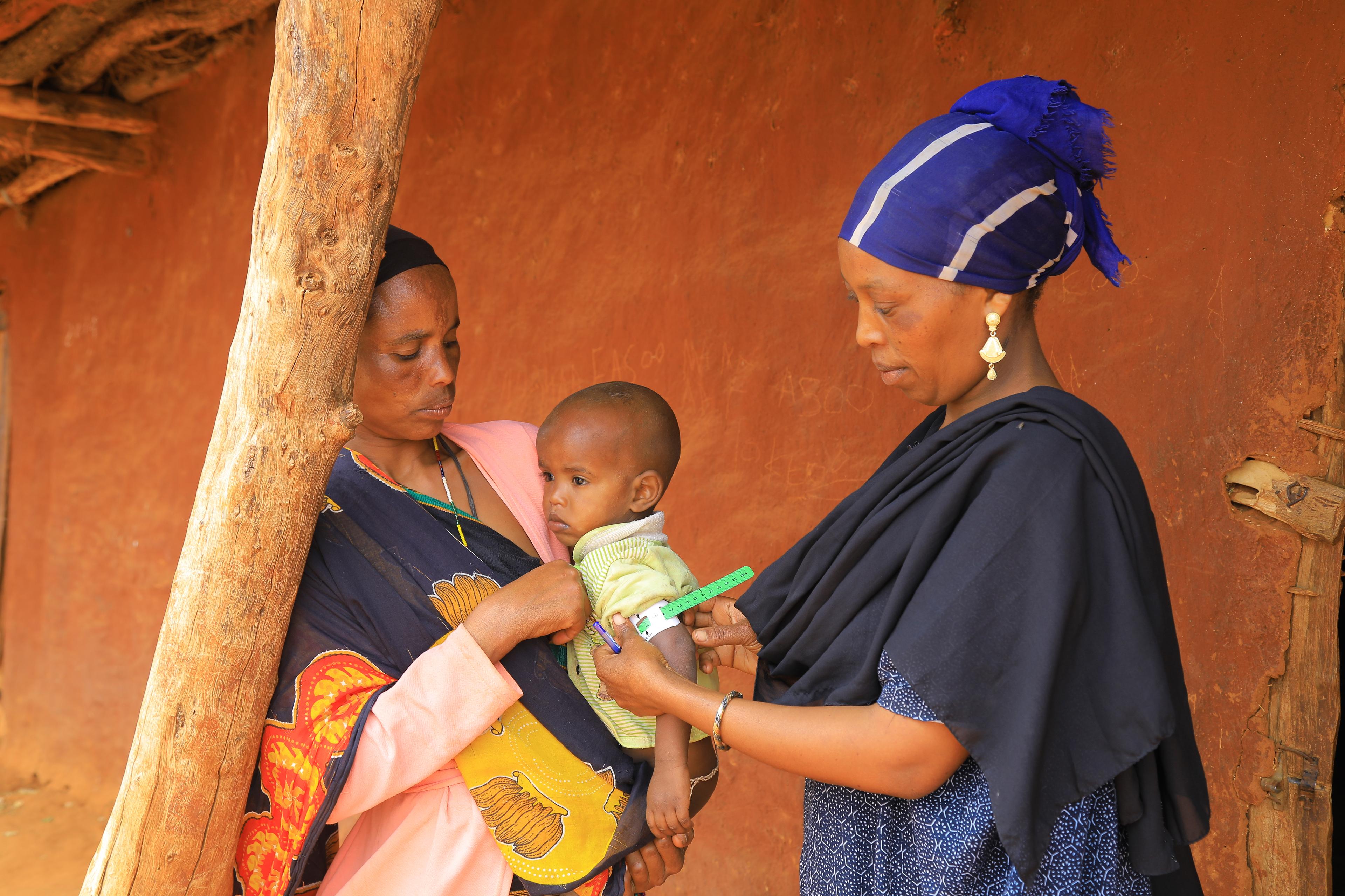 Deux femmes se font face devant une maison en torchis. Celle de gauche porte un bébé dans une écharpe colorée, tandis que celle de droite, vêtue d’une robe et d’un foulard bleus, mesure le tour de bras de l’enfant avec un mètre.