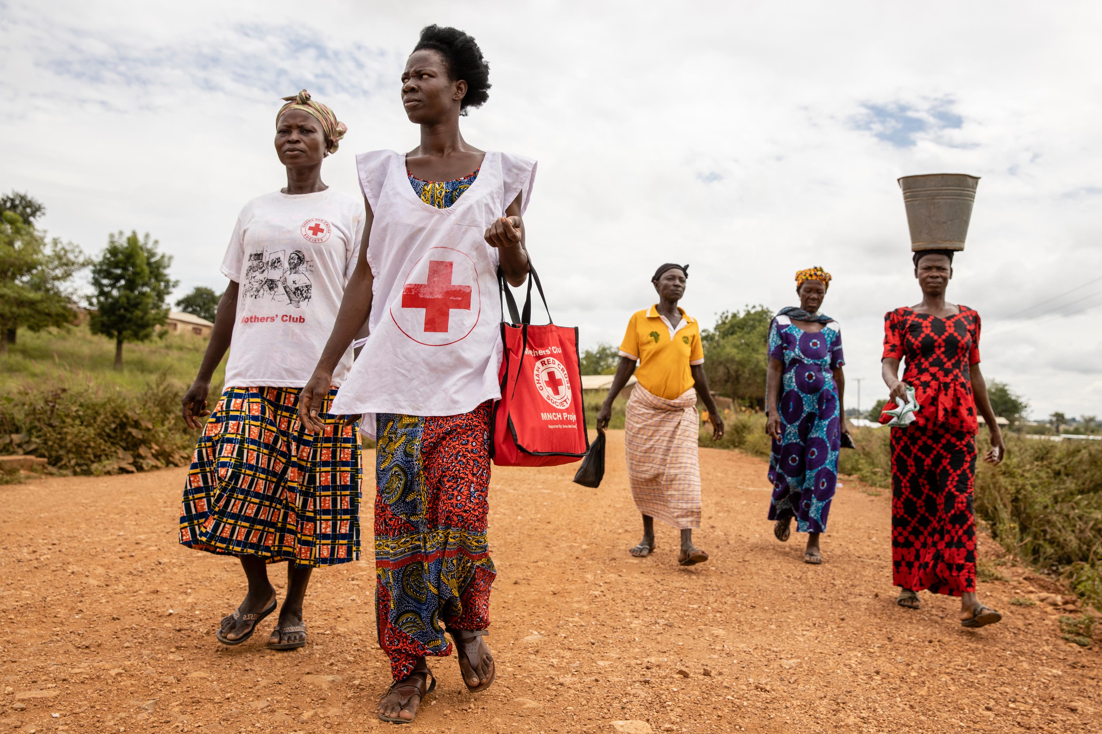 Cinq femmes se déplacent à pied. L'une porte un grand seau sur la tête, deux portent des T-shirts du Mothers Club de la Croix-Rouge ghanéenne.
