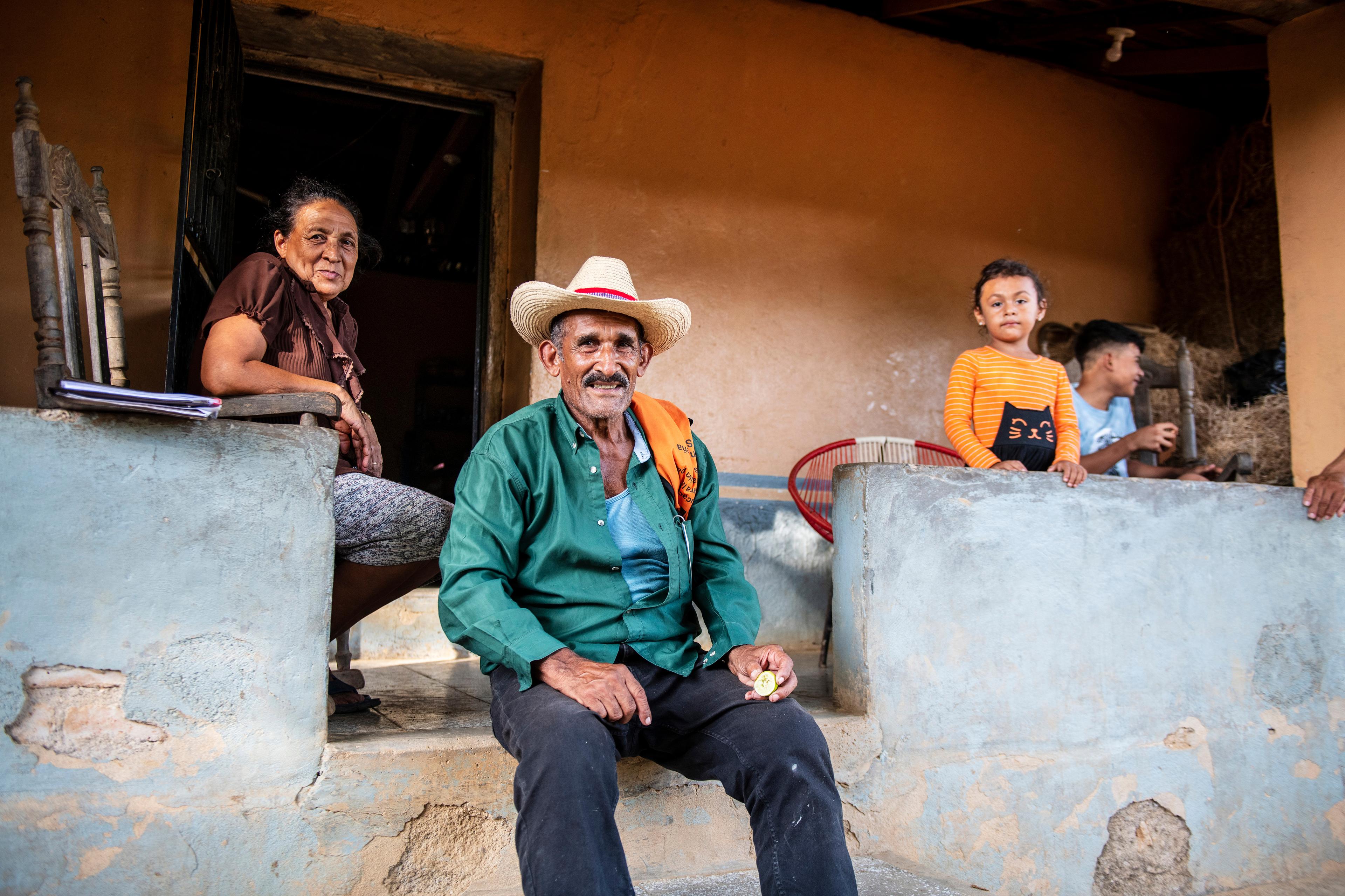 Ein älterer Mann mit Hut und eine Frau sitzen vor einem Haus und lächeln. Daneben stehen zwei Kinder.