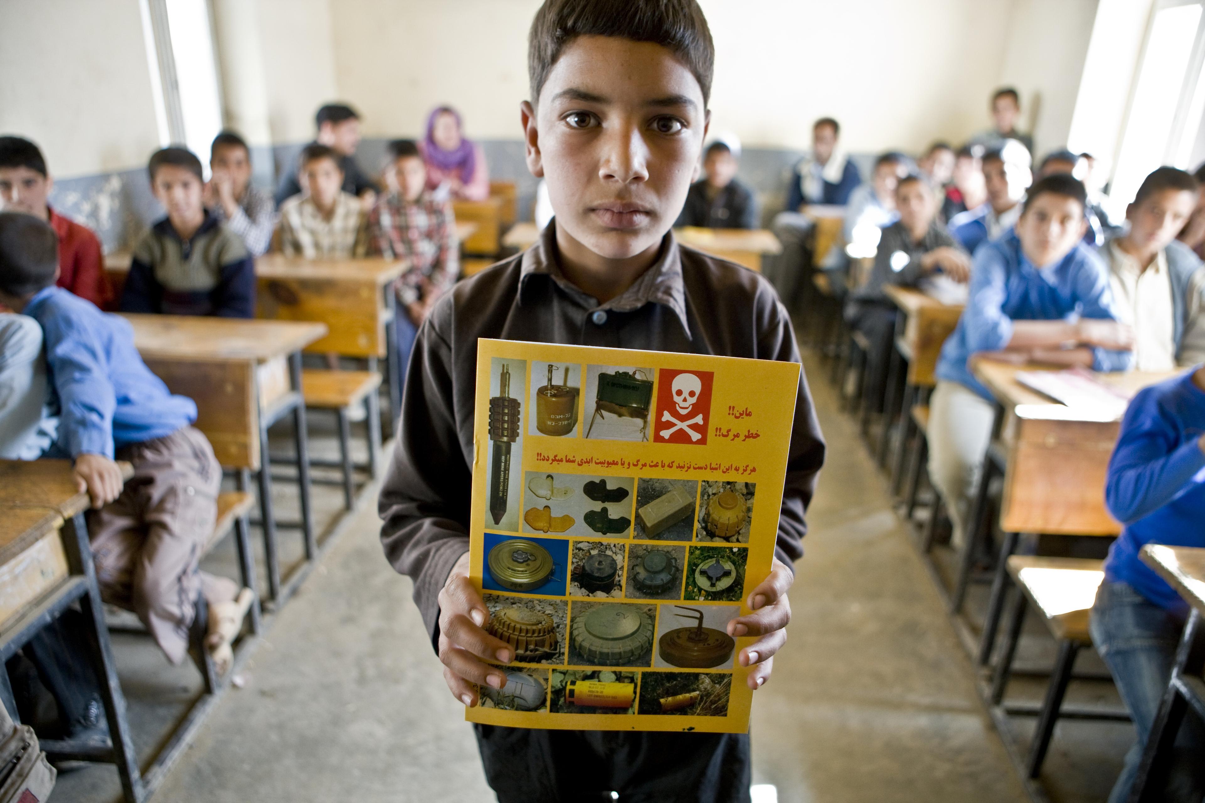 Ein Junge steht im Klassenzimmer und hält ein Heft hoch, das über Minen aufklärt. Seine Mitschüler:innen sind im Hintergrund.