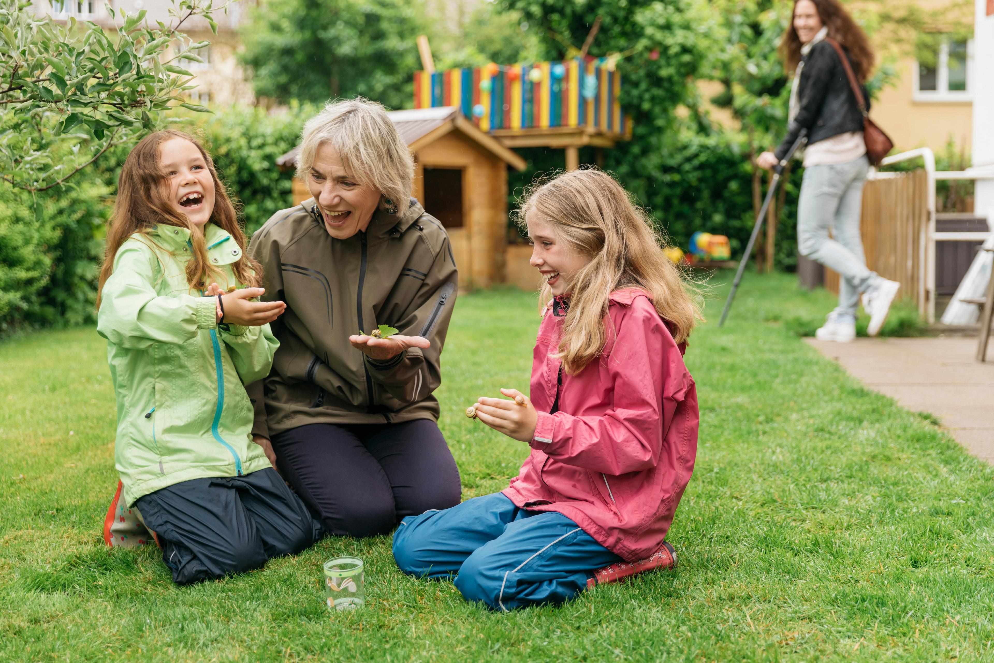 Kinderbetreuuerin und zwei Mädchen sitzen lachend im Garten und halten Schnecken in der Hand. Im Hintergrund ist die Mutter zu sehen. Sie geht an Krücken.