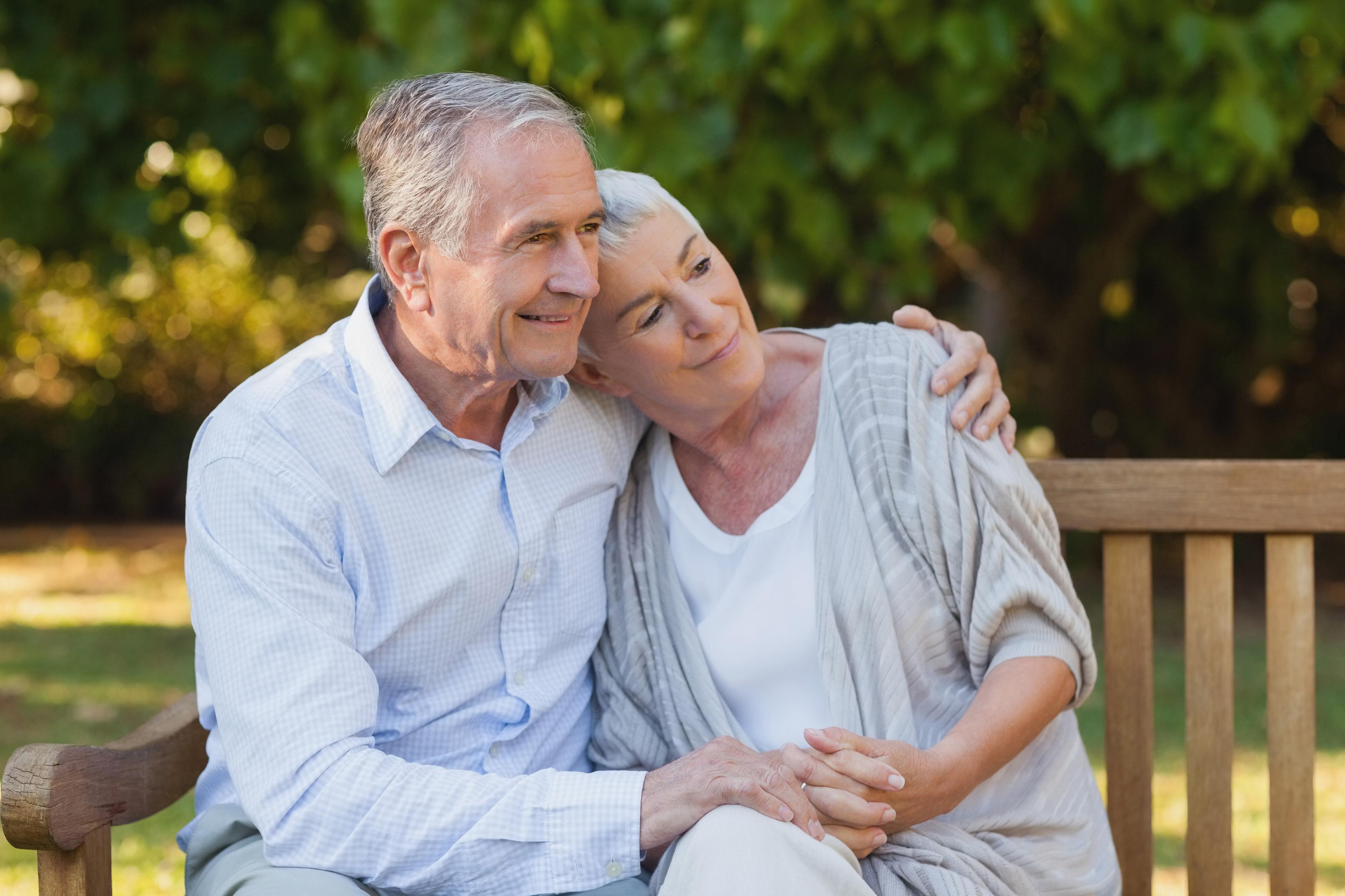 An elderly couple sits on a park bench, holding each other contentedly in their arms.