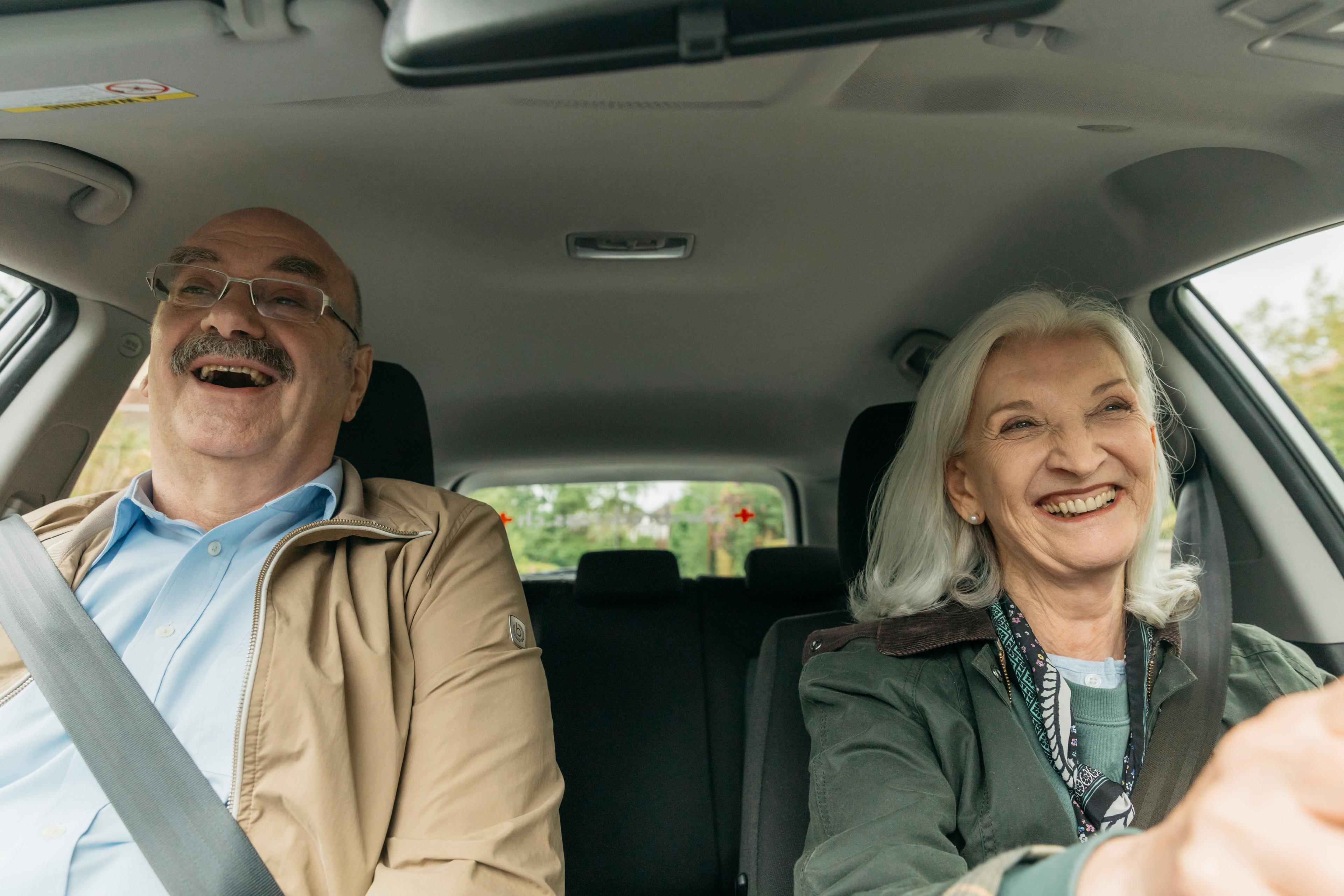 Fahrdienst: Eine ältere Dame sitzt beim Steuerrad und ein älterer Herr sitzt auf dem Beifahrersitz. Beide lachen.