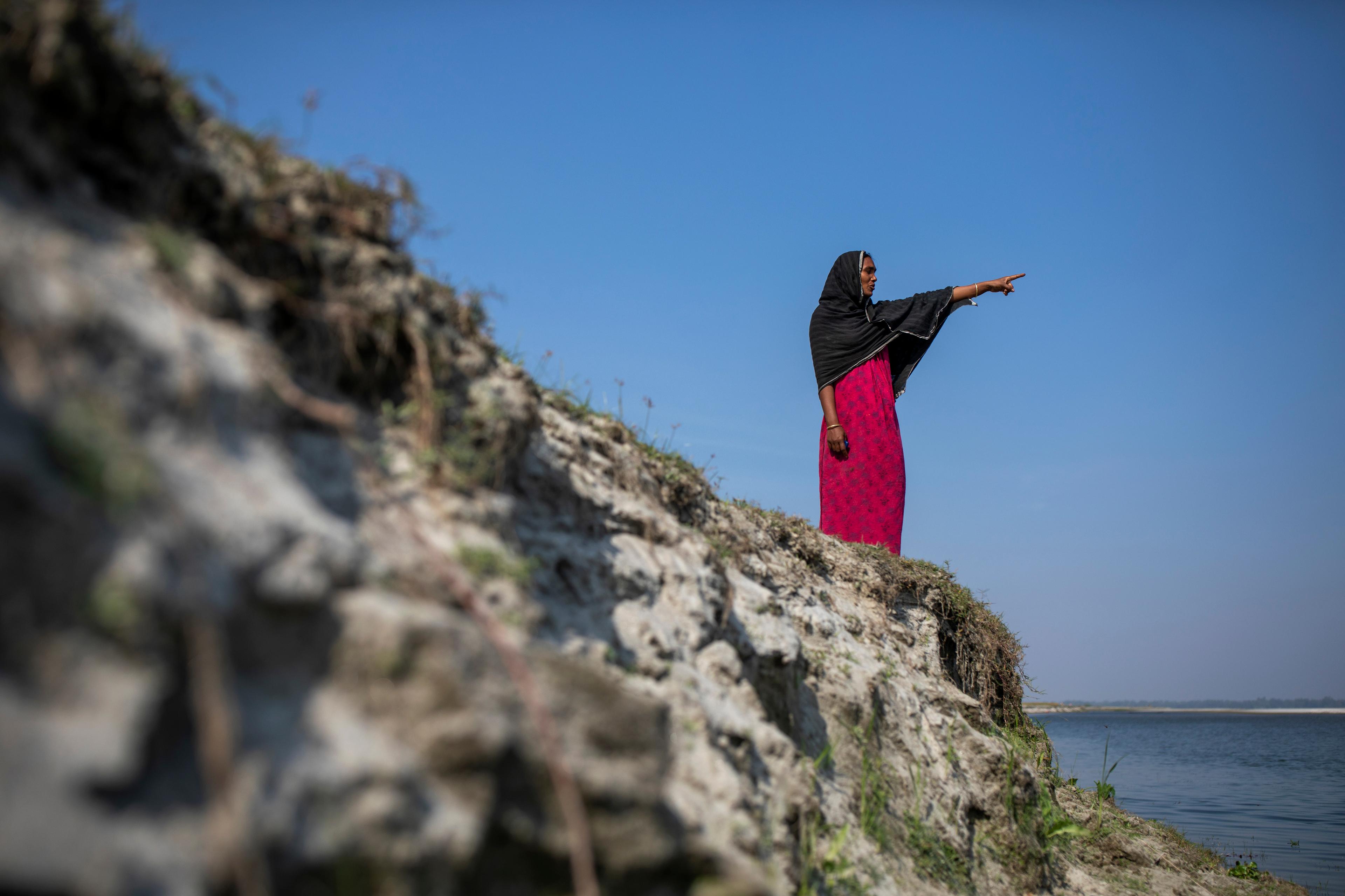 Una donna indica in lontananza con il braccio sinistro. Si guarda la mano. Tutto il suo corpo è visibile, è in piedi vicino a un fiume. Il cielo sullo sfondo è blu.