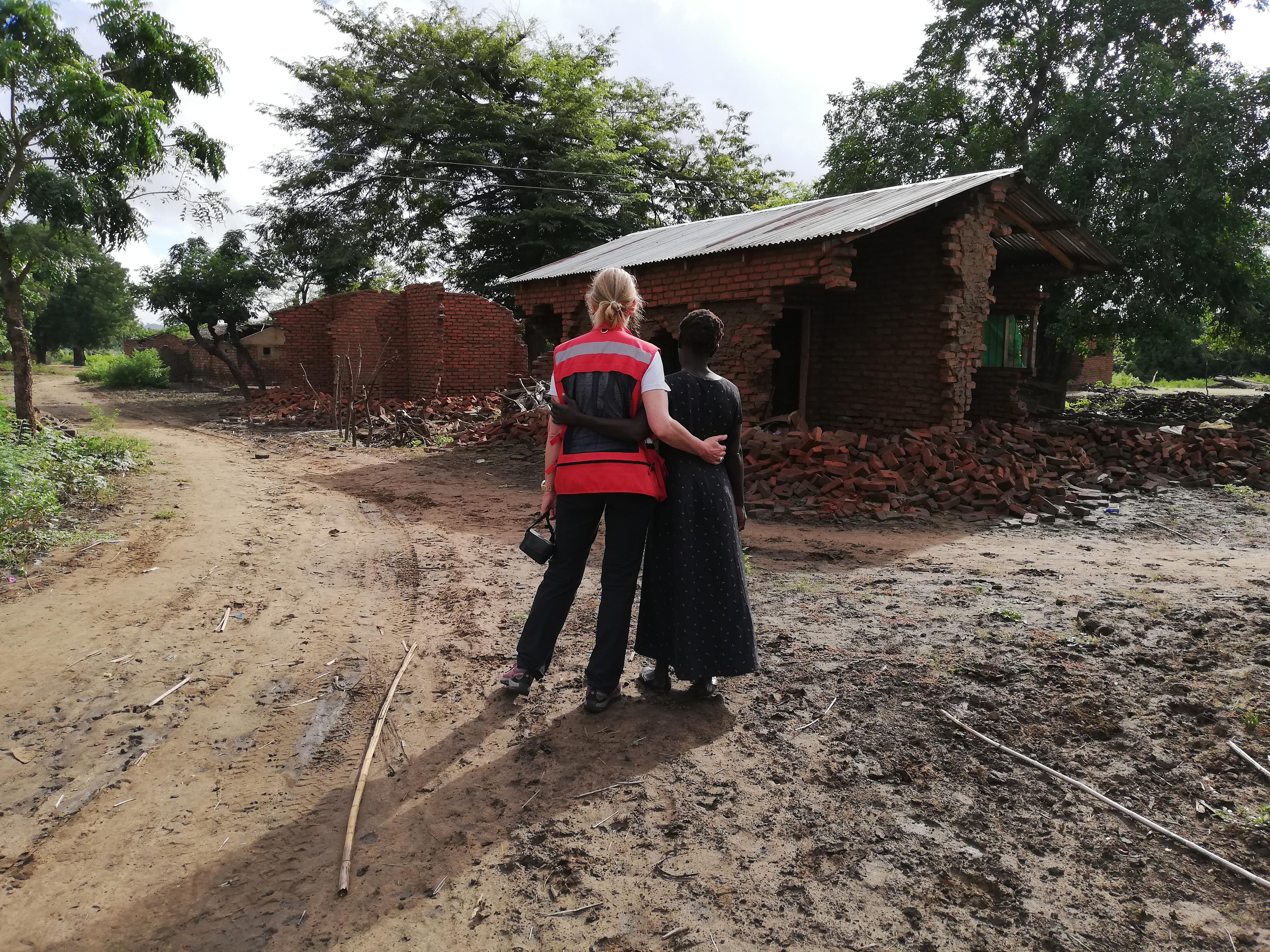Red Cross delegate Regina Wenk holds a woman from the village of Marica in her arms; they are standing in front of a destroyed house.
