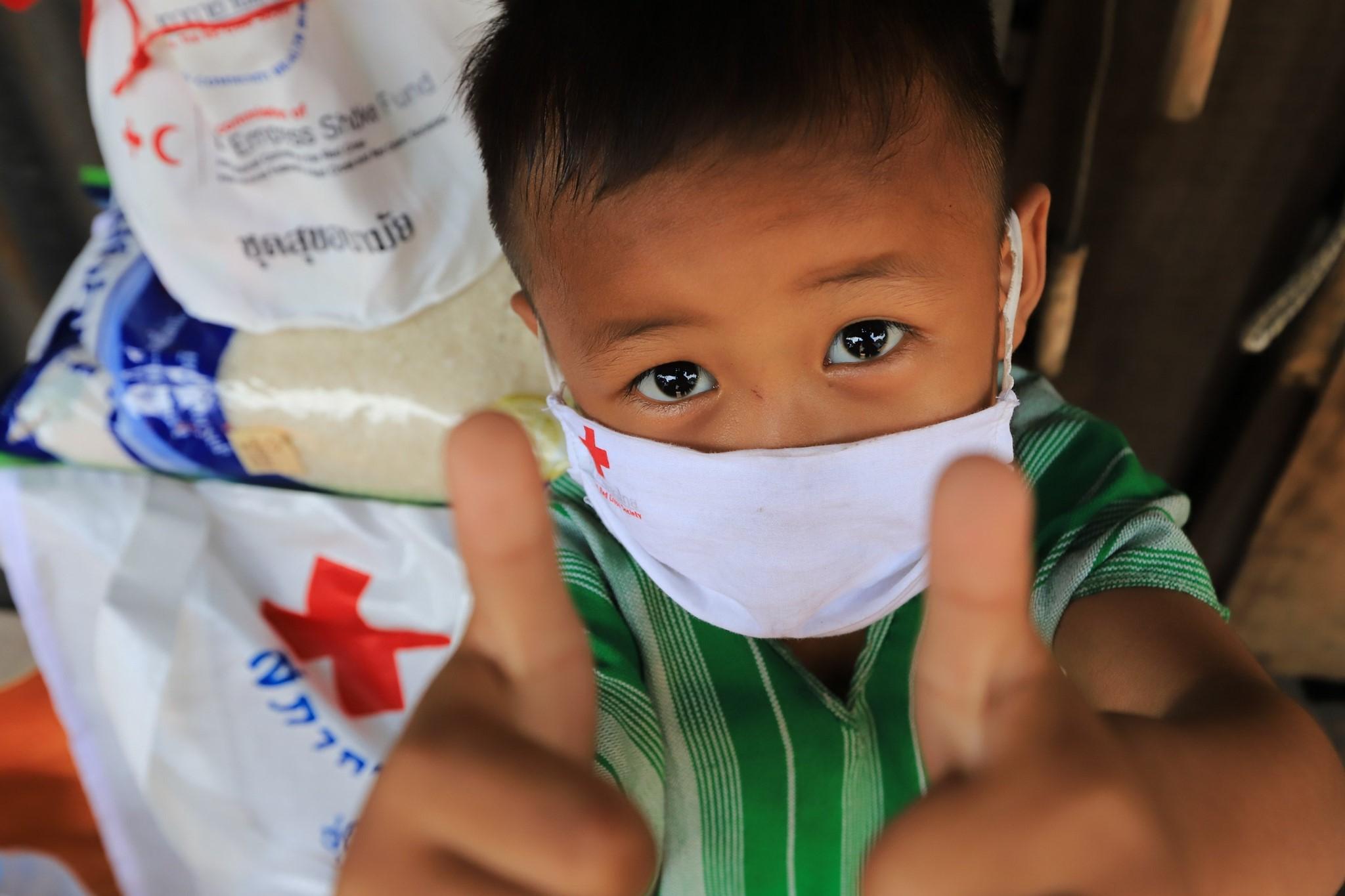 Ein kleiner Junge lächelt hinter seiner Maske mit dem Logo des Roten Kreuzes. Er hält beide Daumen hoch. Im Hintergrund steht eine Ladung Reis mit dem Emblem des Roten Kreuzes auf dem Tisch.  