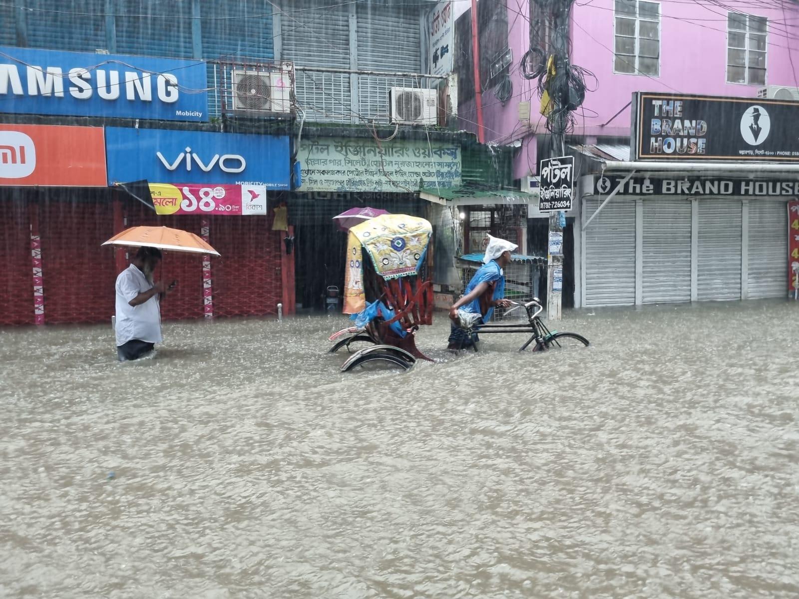Un homme tire son rickshaw sous une pluie battante dans une rue inondée où les commerces sont fermés.  Derrière lui, un homme marche avec un parapluie. Ils ont de l'eau jusqu'à la taille.