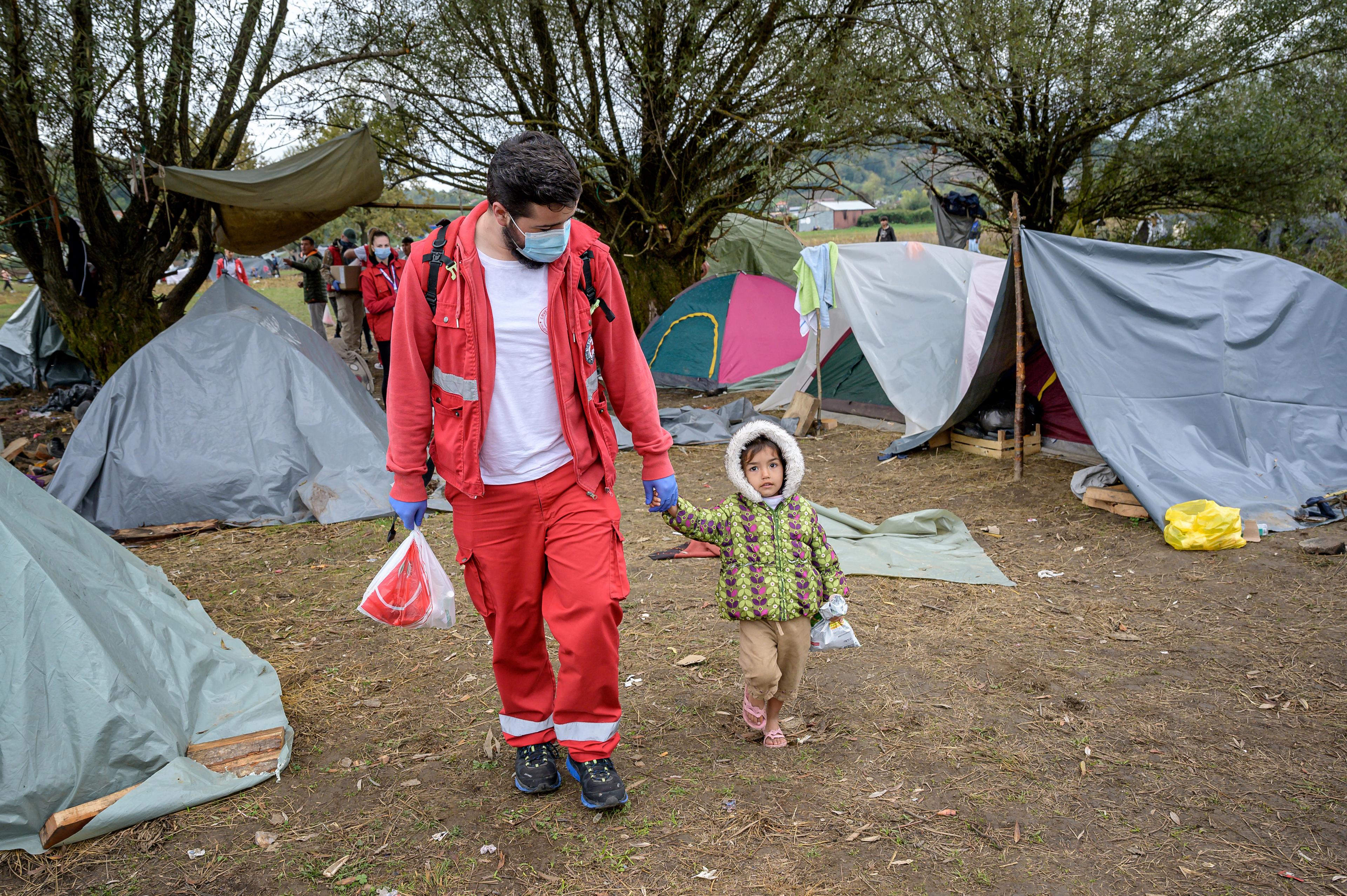 A man wearing red pants and a red jacket is holding a small child with his left hand. They walk together through tents, which are located on a green area. 
