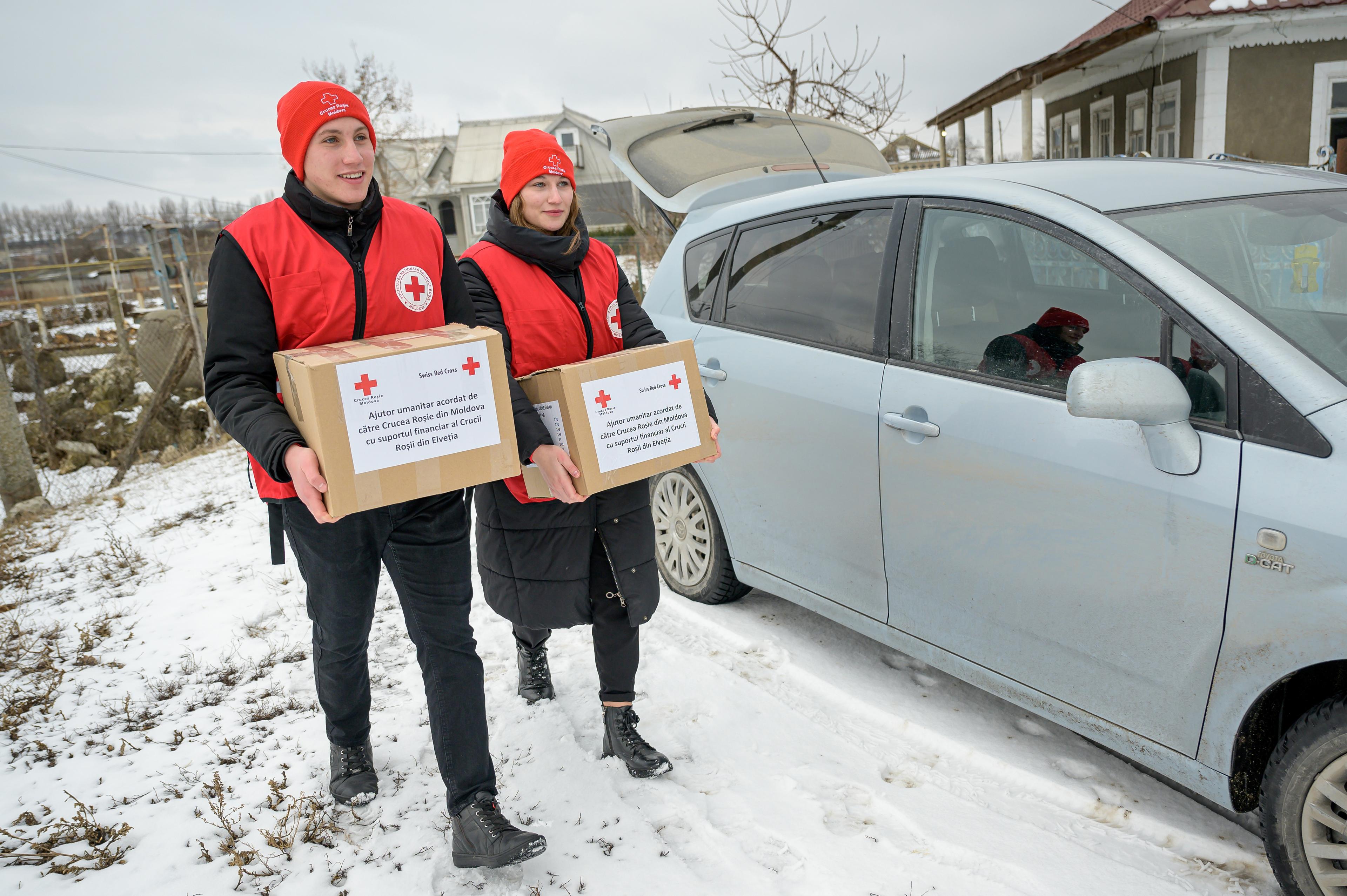 Deux jeunes adultes tiennent un paquet dans leurs mains et marchent à côté d'une voiture. Ils sont habillés de vêtements épais et portent un gilet et un bonnet rouges. Il y a de la neige sur le sol.