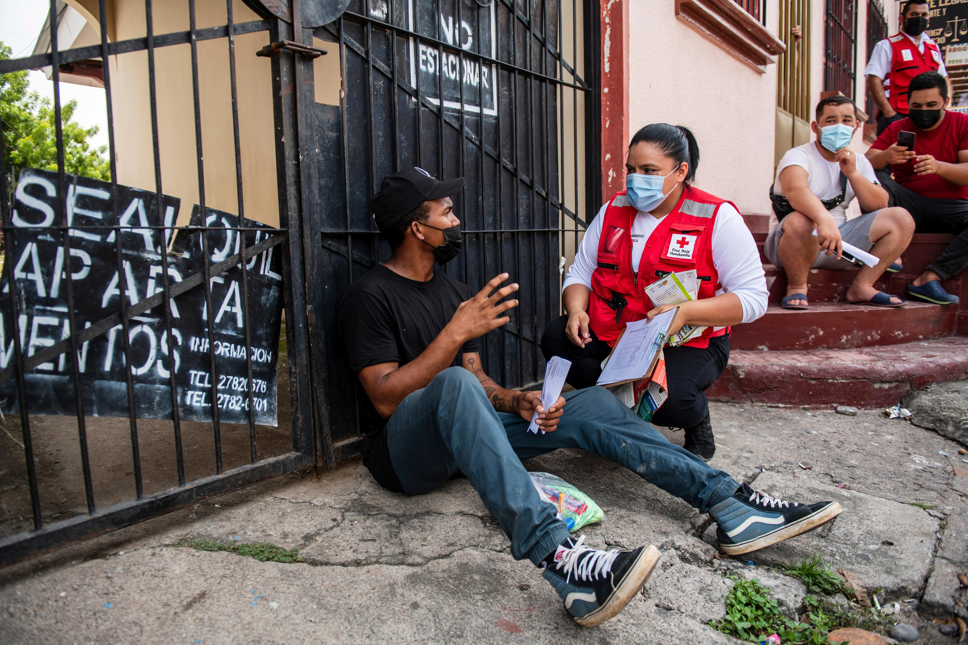 A woman is wearing a red vest and a hygiene mask. She kneels next to a man sitting on the floor and talks to him. In her hand she holds documents. They are in front of a gate. More men are sitting in the background. 