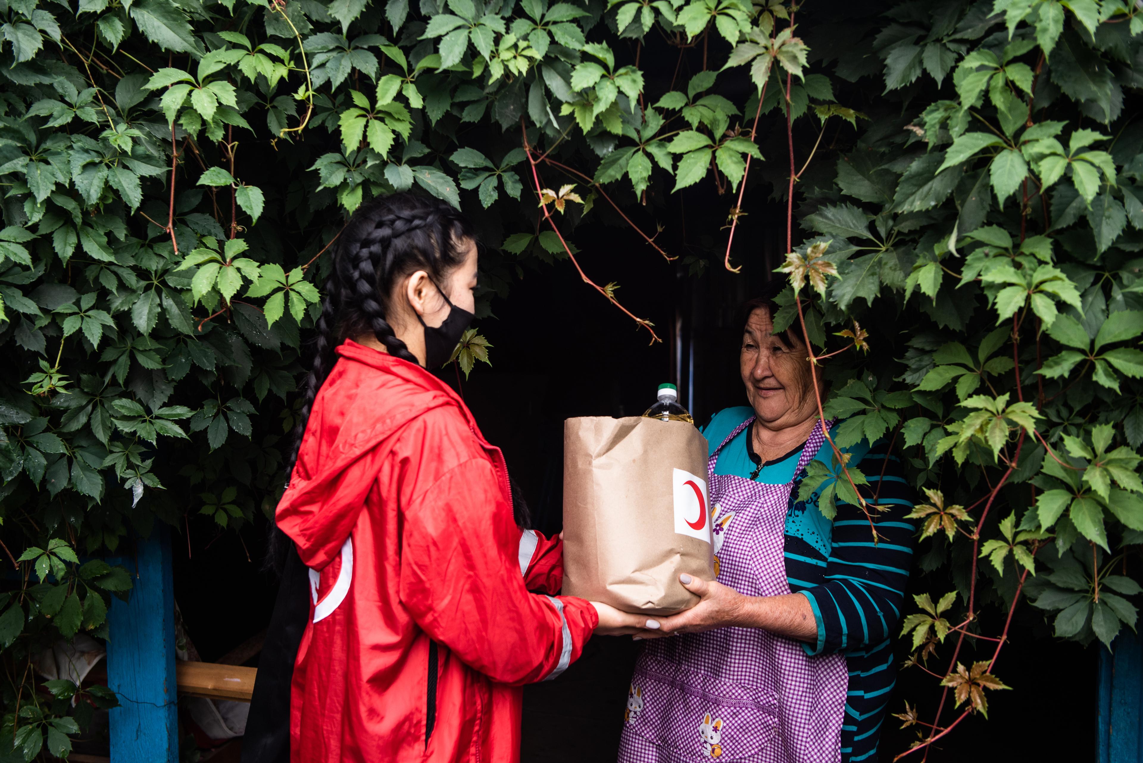 A volunteer of the Kyrgyz Red Crescent, hands a food package to beneficiary Nurmukhanbetova Karlygash. She is standing in front of a house heavily covered with plants.