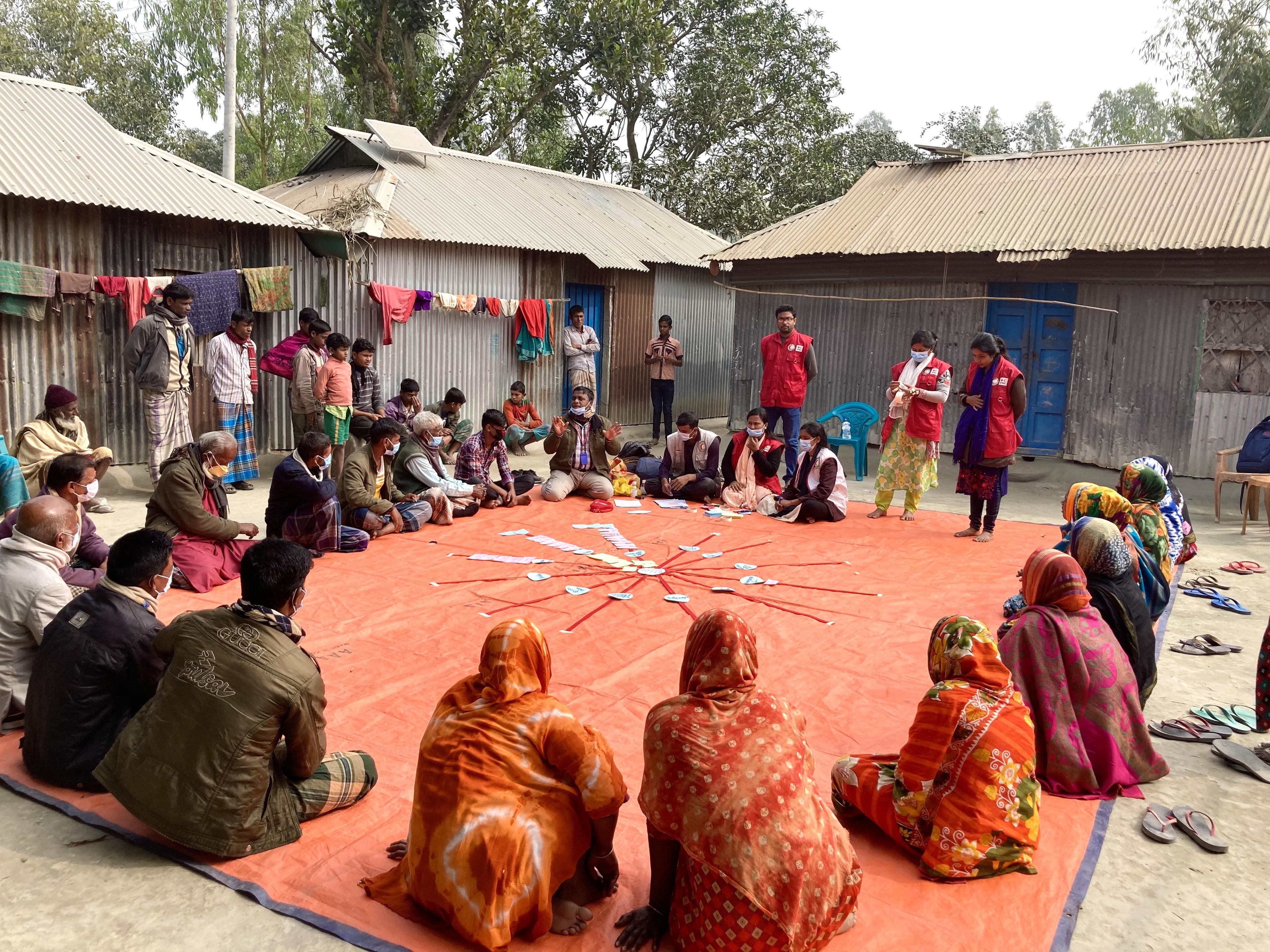 Women and men sit in a circle on the ground in a village in Bangladesh. They have laid out labeled cards in the center. Volunteers from the Bangladesh Red Crescent can also be seen.