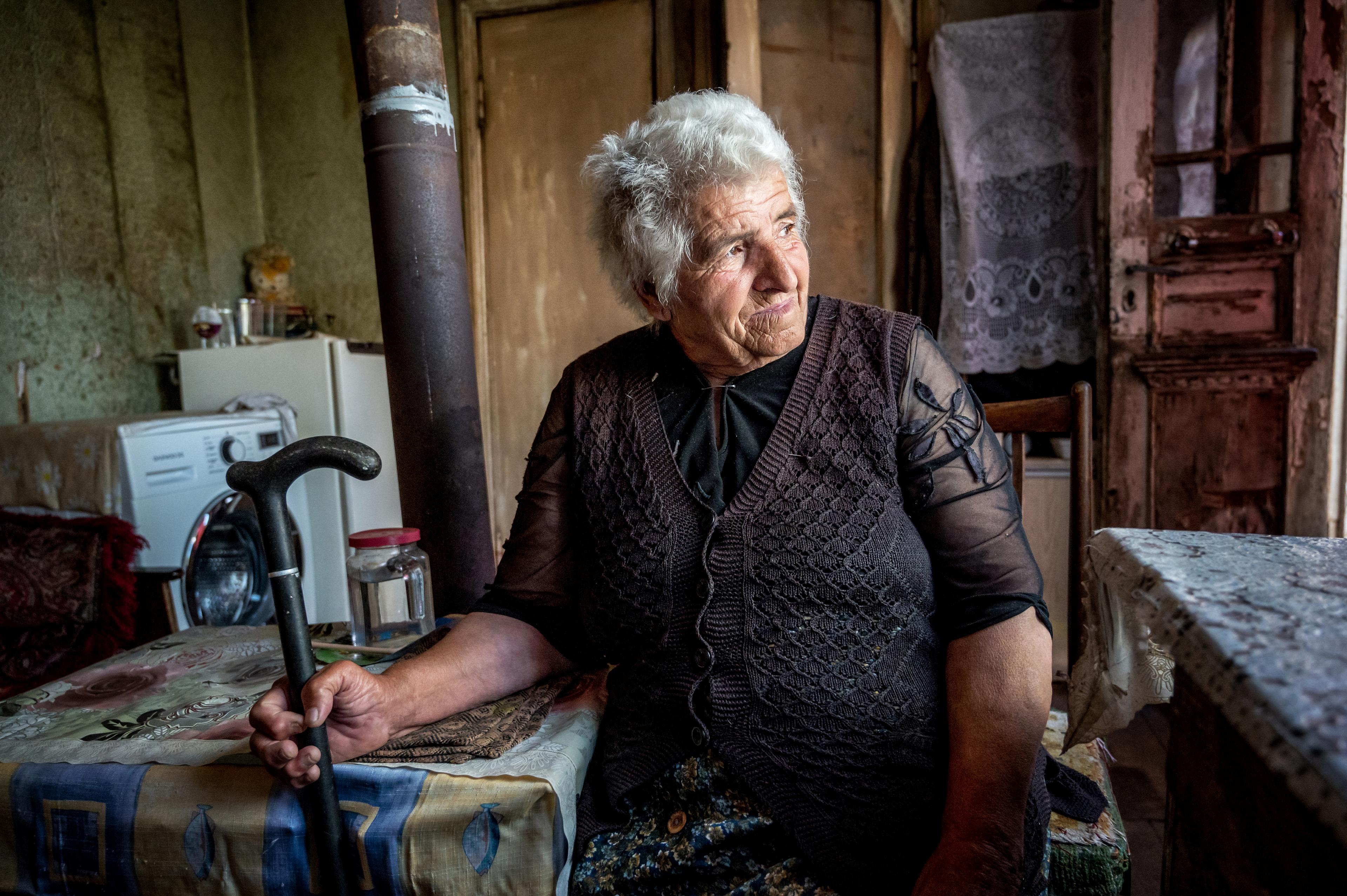 Une femme âgée est assise à une table et regarde pensivement par la fenêtre. Elle tient une canne dans sa main droite.