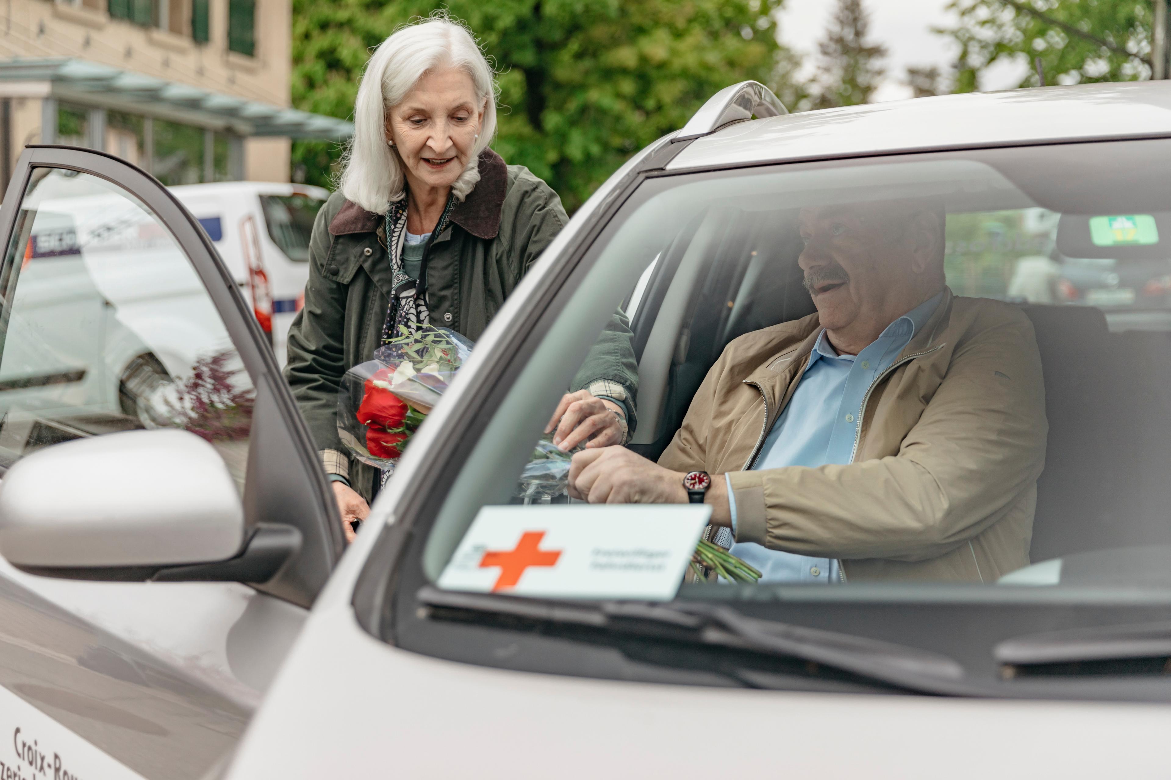 Service des transports: une femme aide un homme âgé à monter dans une voiture. Elle lui tend un bouquet de fleurs.