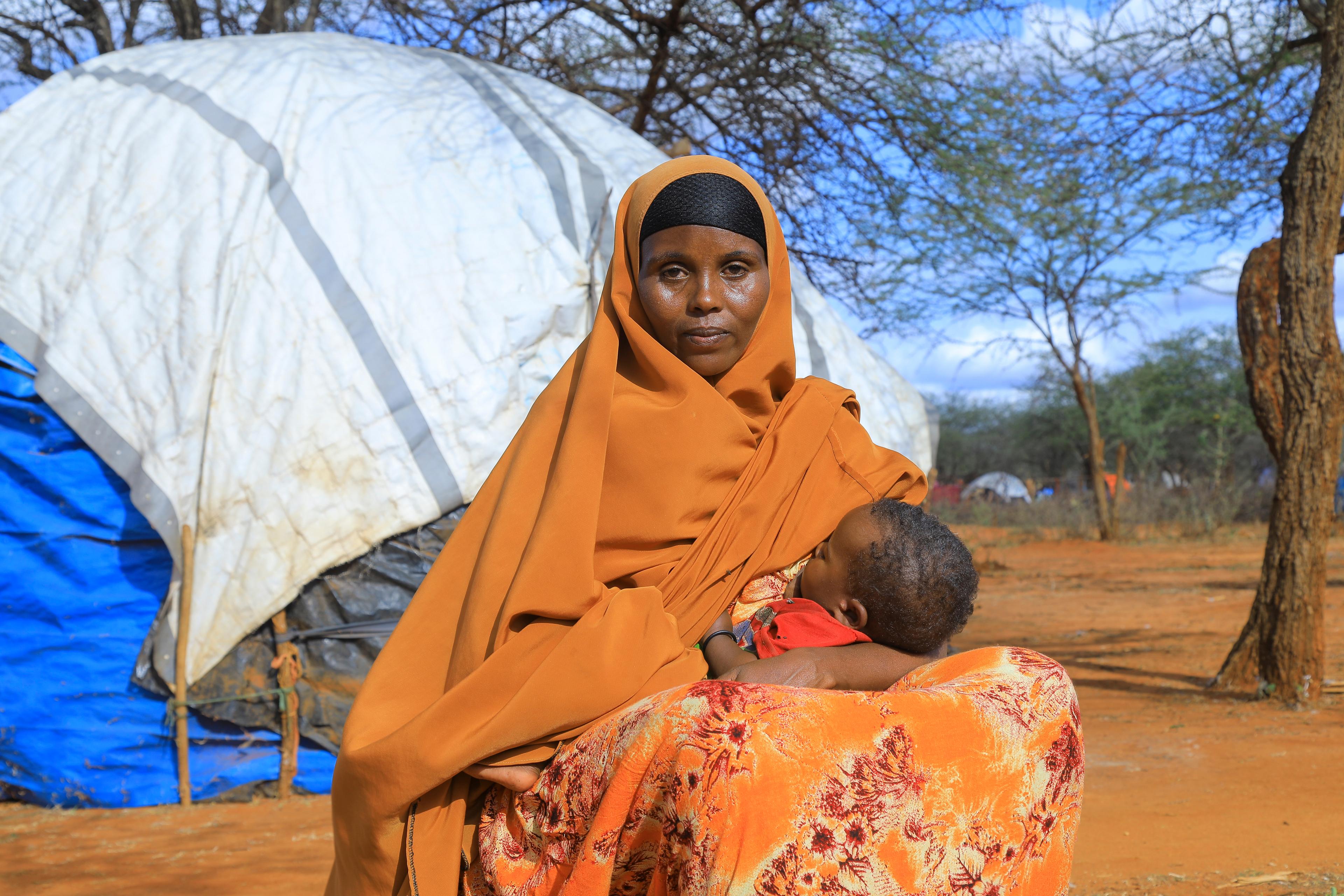 Una donna con un velo arancione e un abito dello stesso colore allatta il suo bambino davanti a una tenda.