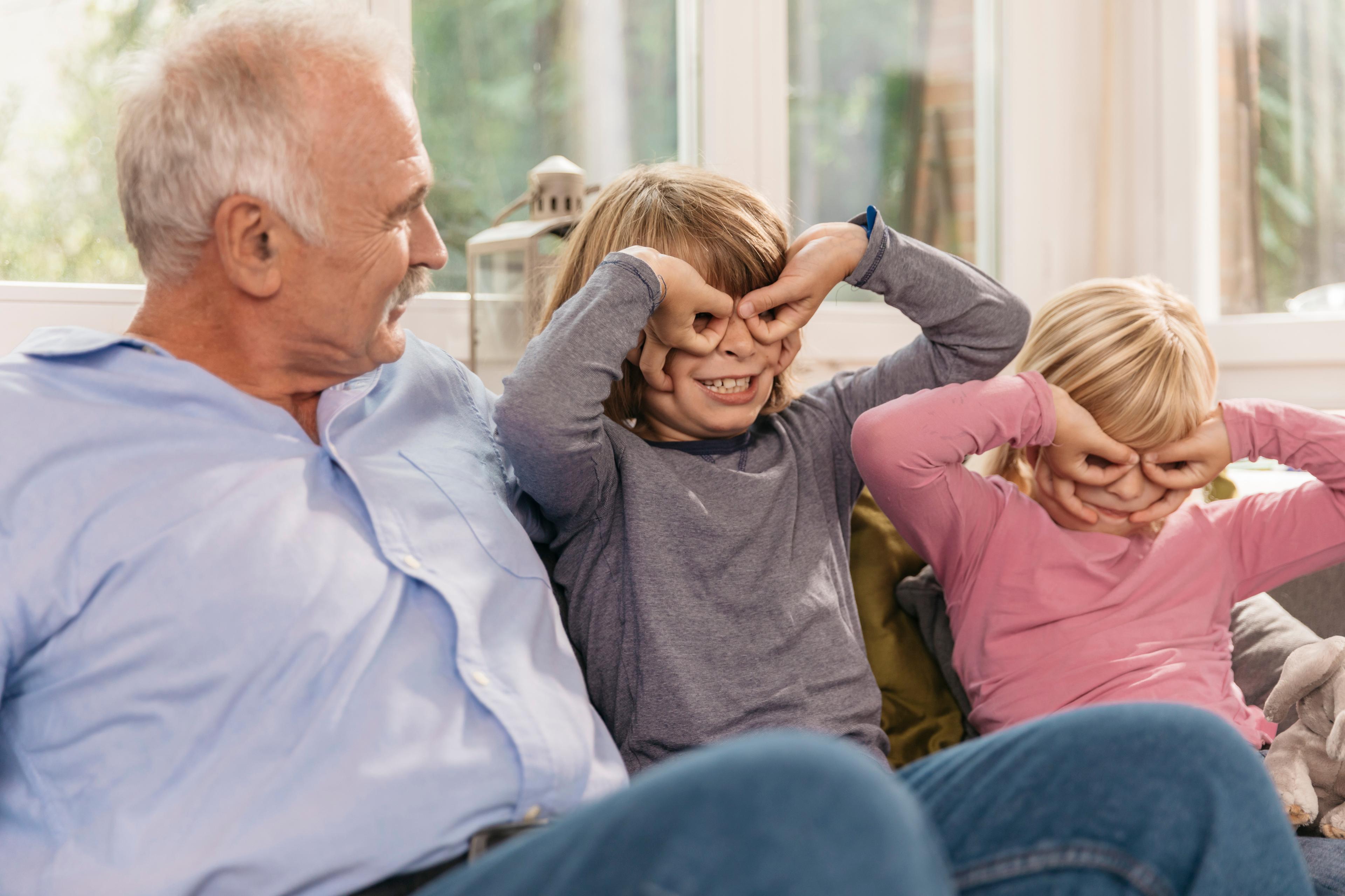 Due bambini stanno giocando a mimare gli occhiali con le mani davanti agli occhi. I bambini sono seduti su un divano con il nonno. Stanno tutti ridendo.