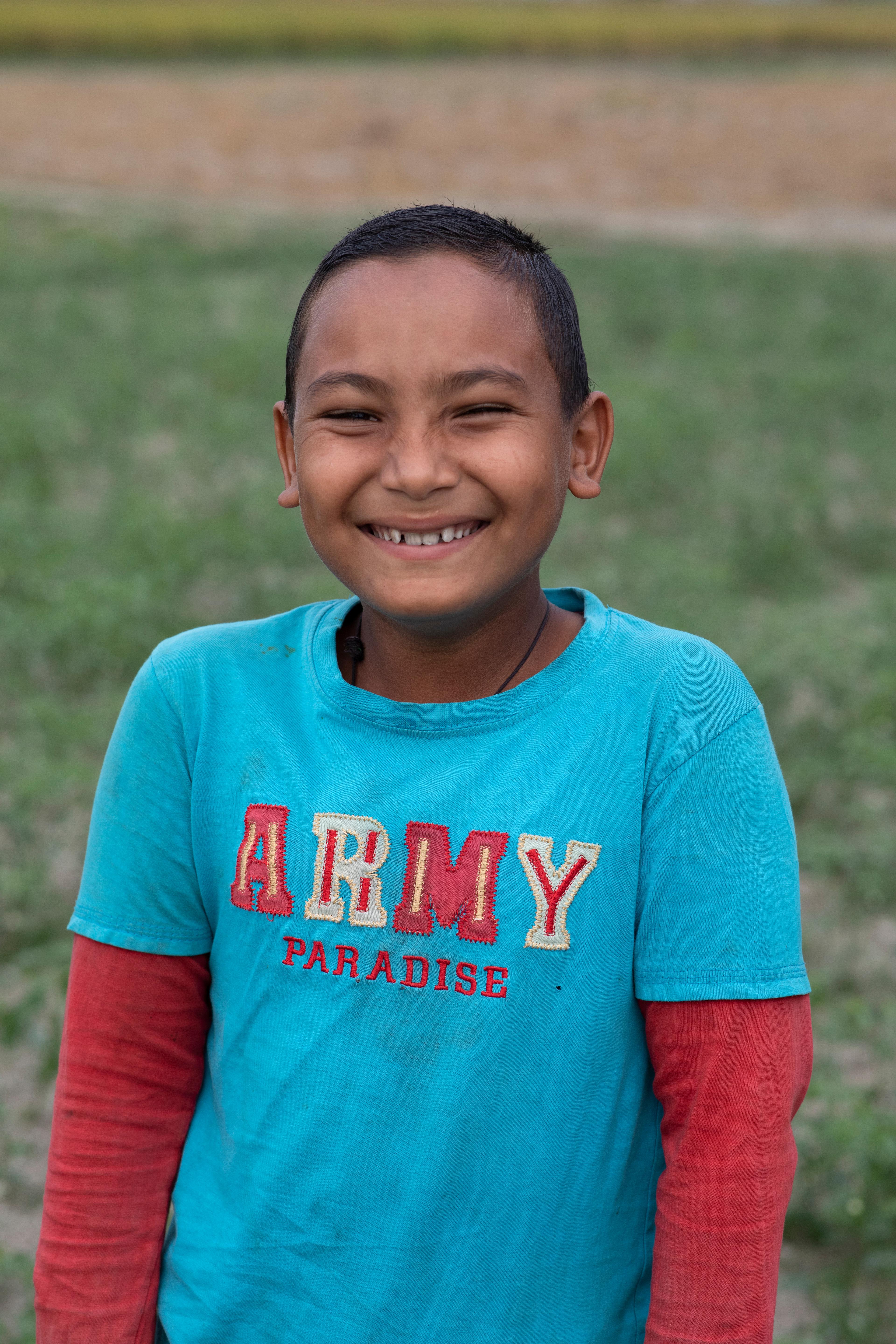 Portrait eines Jungen. Er trägt ein breites Lächeln im Gesicht und ein T-Shirt über einem Pullover. Im Hintergrund ist ein durch die Kamera verschwommenes Feld.