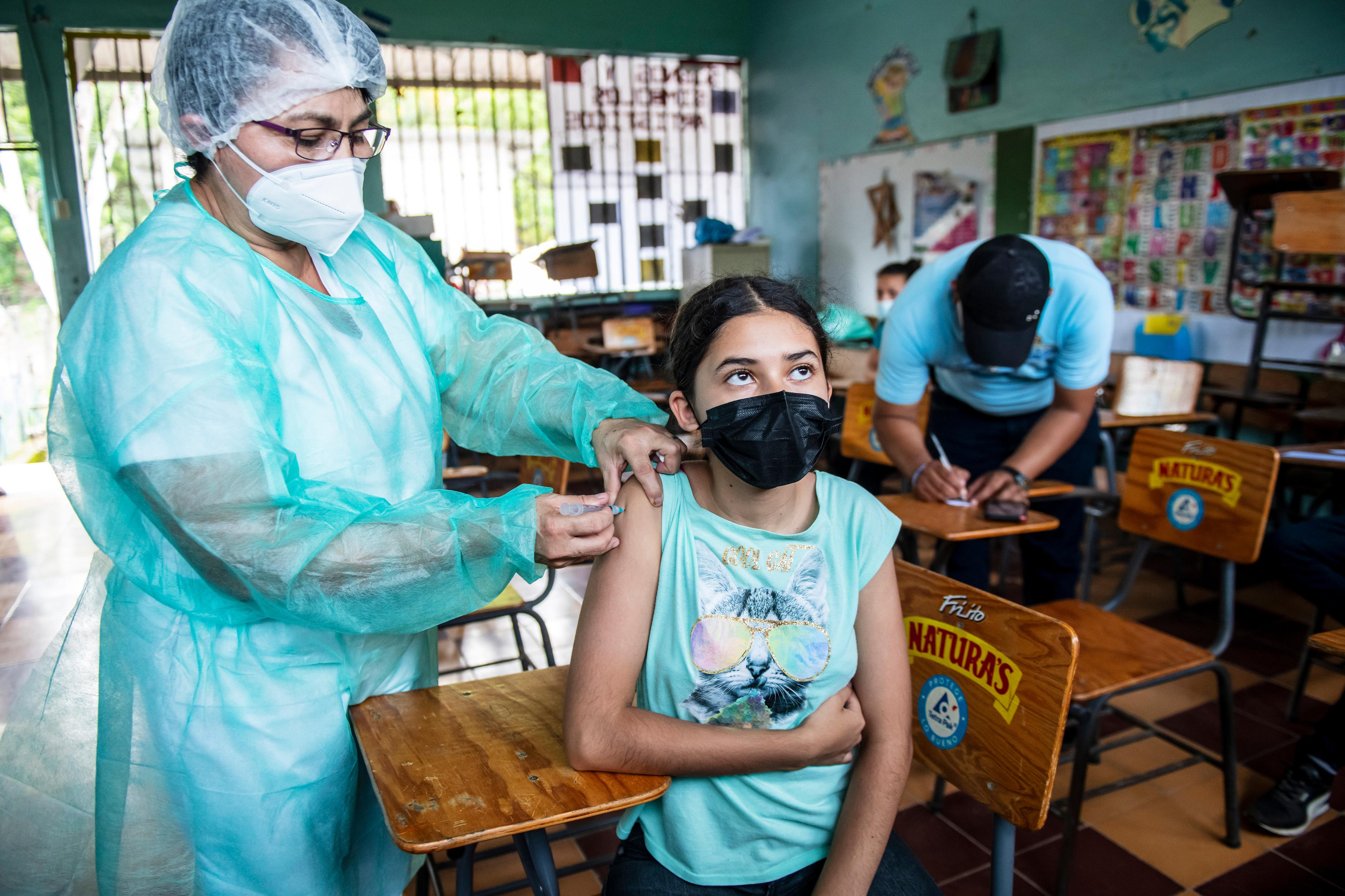 Una donna in abbigliamento protettivo vaccina un adolescente. Si trovano in un edificio scolastico.