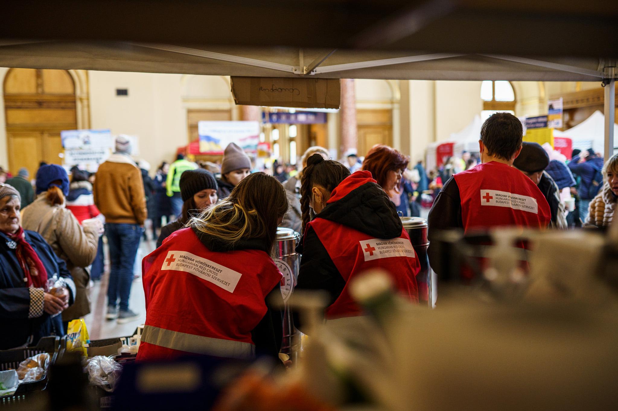 Nella foto ci sono tre giovani di spalle, tutti indossano un gilet rosso con il simbolo della Croce Rossa. Si trovano in una tenda all’interno di una stazione insieme ad altre persone. Sono impegnati a distribuire acqua e pacchi alimentari.