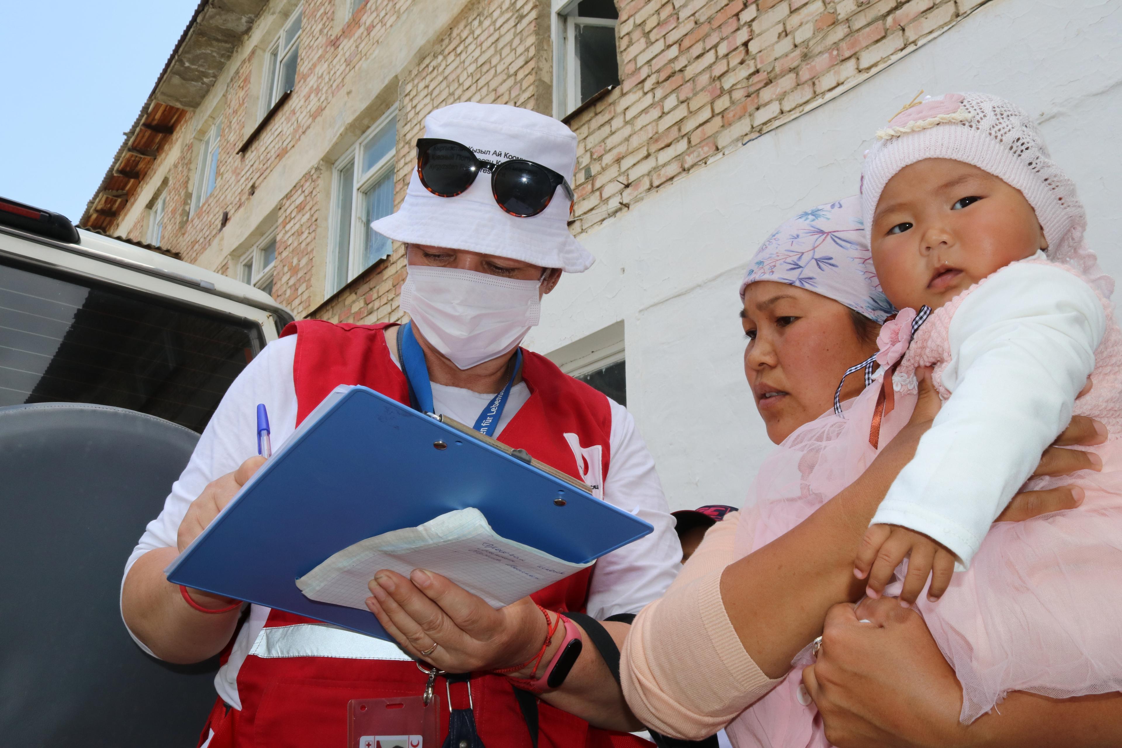 A woman market with Red Cross waistcoat wearing a sanitary mask writingon a sheet and a woman with child in her arms at her right looking at the sheet.