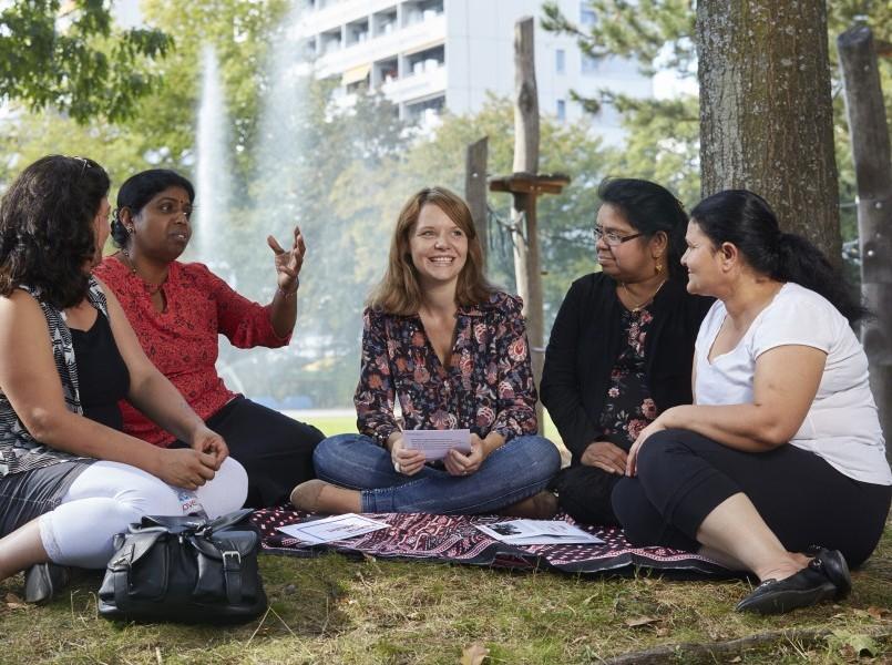 Cinq femmes sont assises en demi-cercle sur une couverture dans un parc municipal, devant un arbre, et discutent.