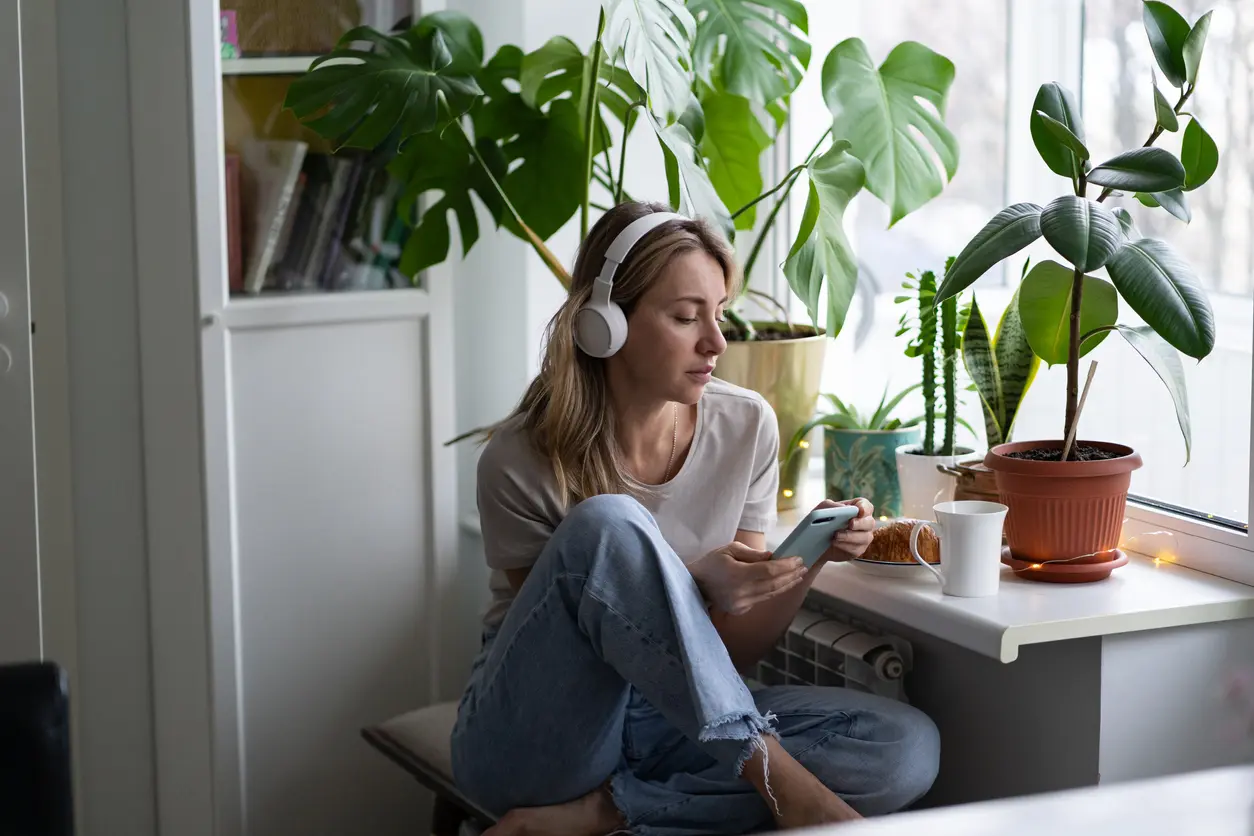 Eine Frau in einer gemütlichen Umgebung Zuhause. Sie hört einen Podcast, trägt drahtlose weisse Kopfhörer und benutzt ein Smartphone.