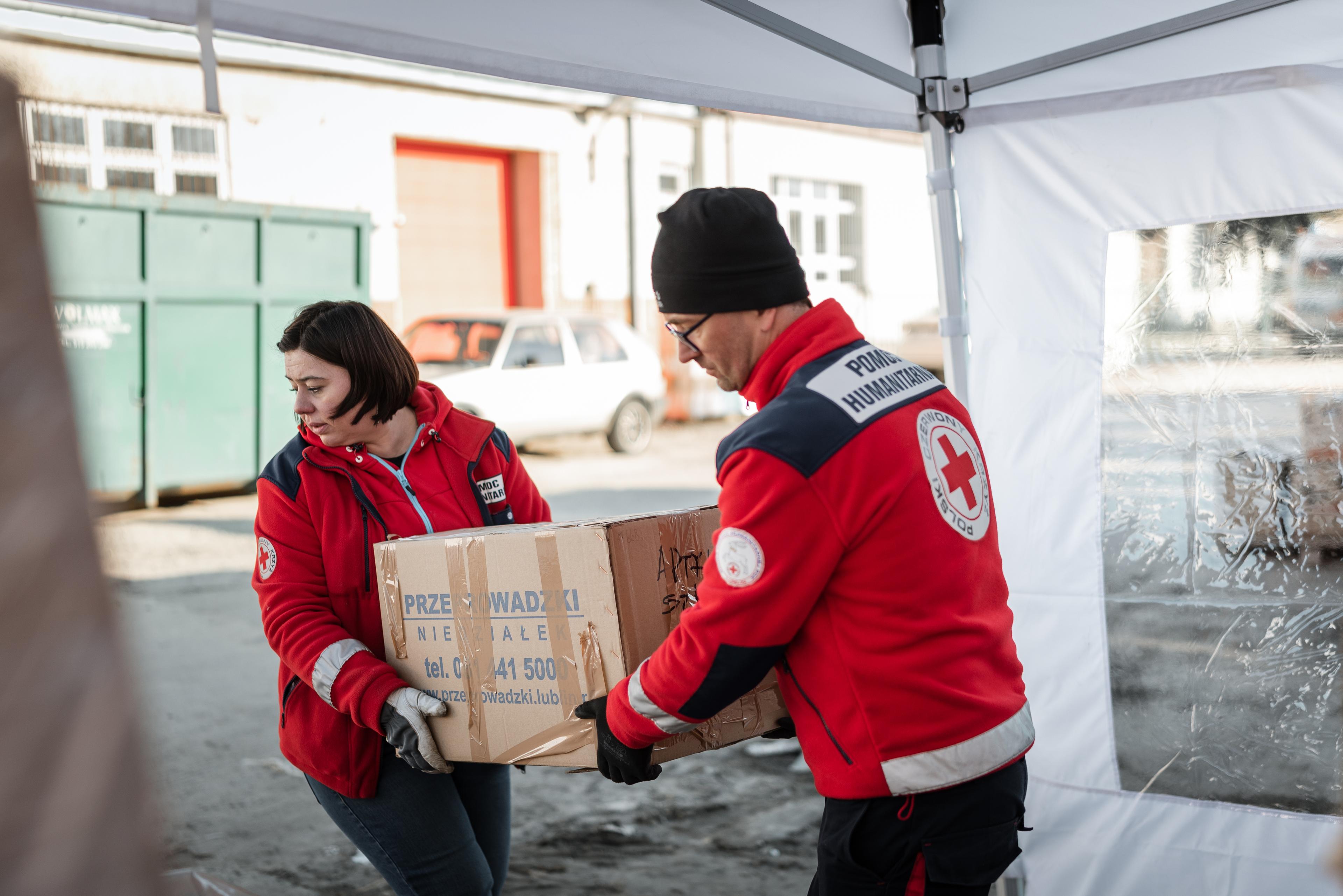 Une bénévole et un bénévole de la Croix-Rouge polonaise portent ensemble une caisse en carton. Il s'agit probablement de matériel de secours.
