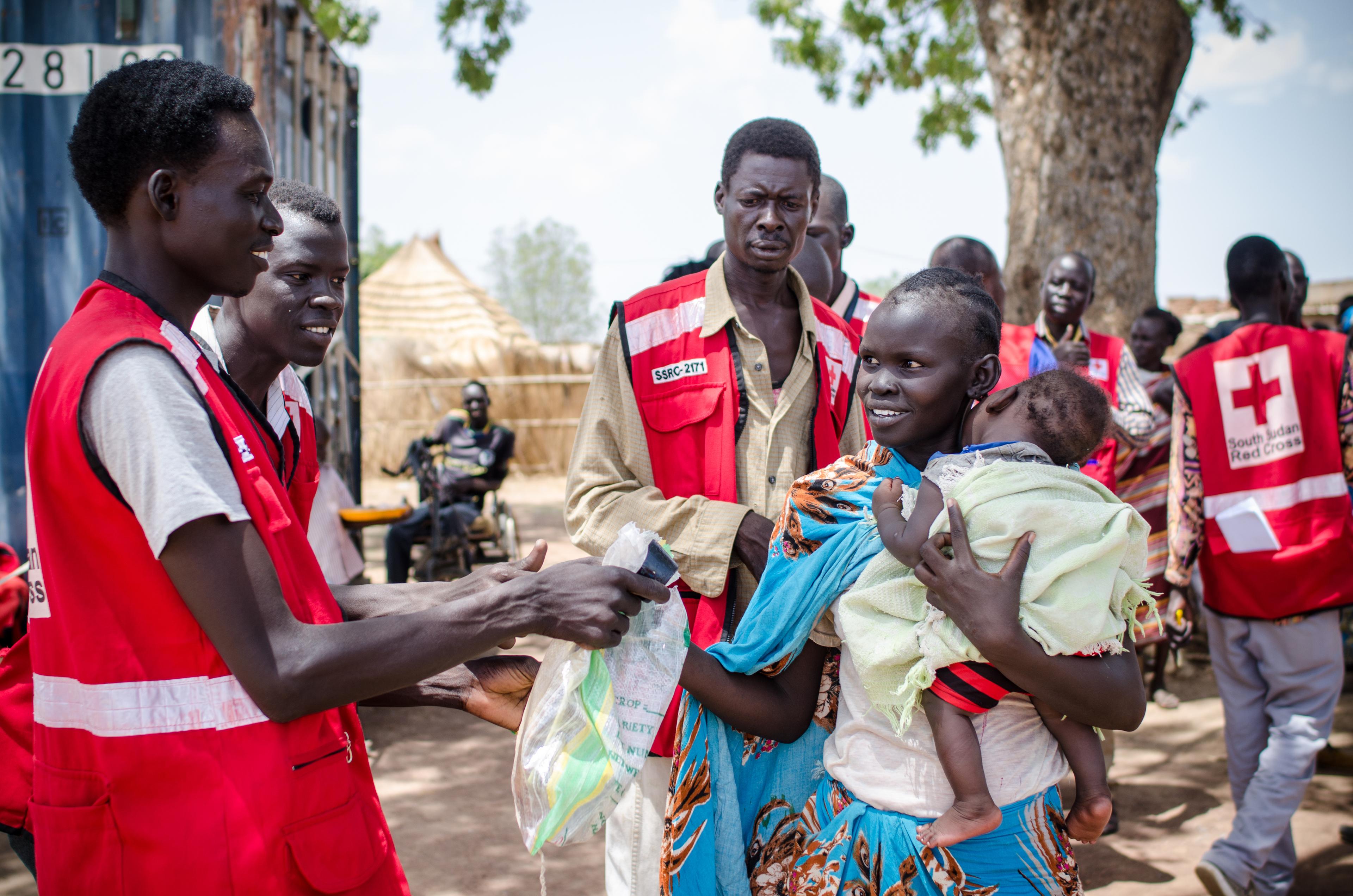 Una donna riceve sementi e attrezzi agricoli in un punto di distribuzione a Panemeth, in Sud Sudan. Porta in braccio un bambino piccolo.
