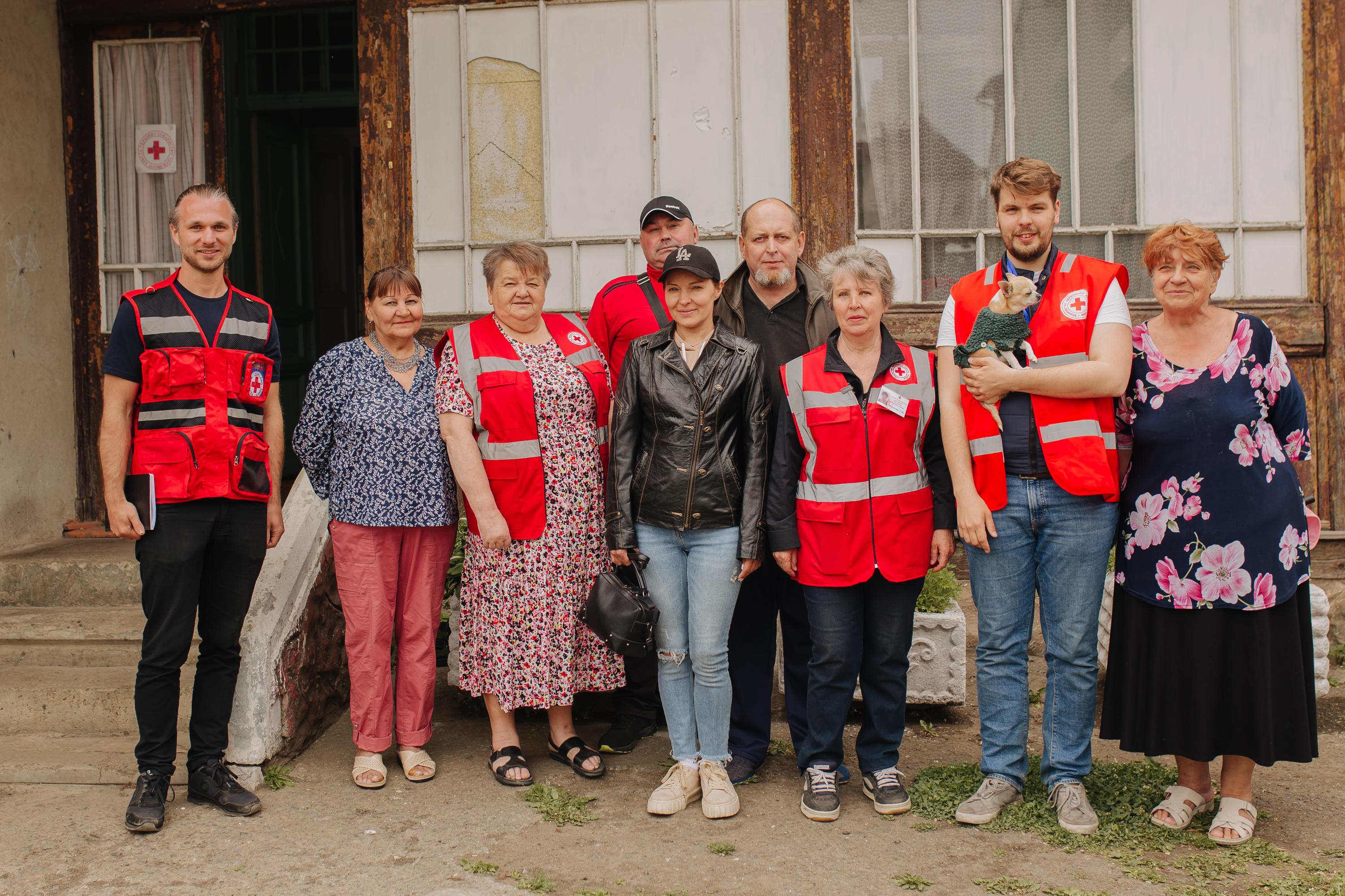 Una foto di gruppo in cui si vedono nove persone. Un giovane uomo tiene in braccio un cane. Quattro persone indossano un gilet rosso della Croce Rossa. 